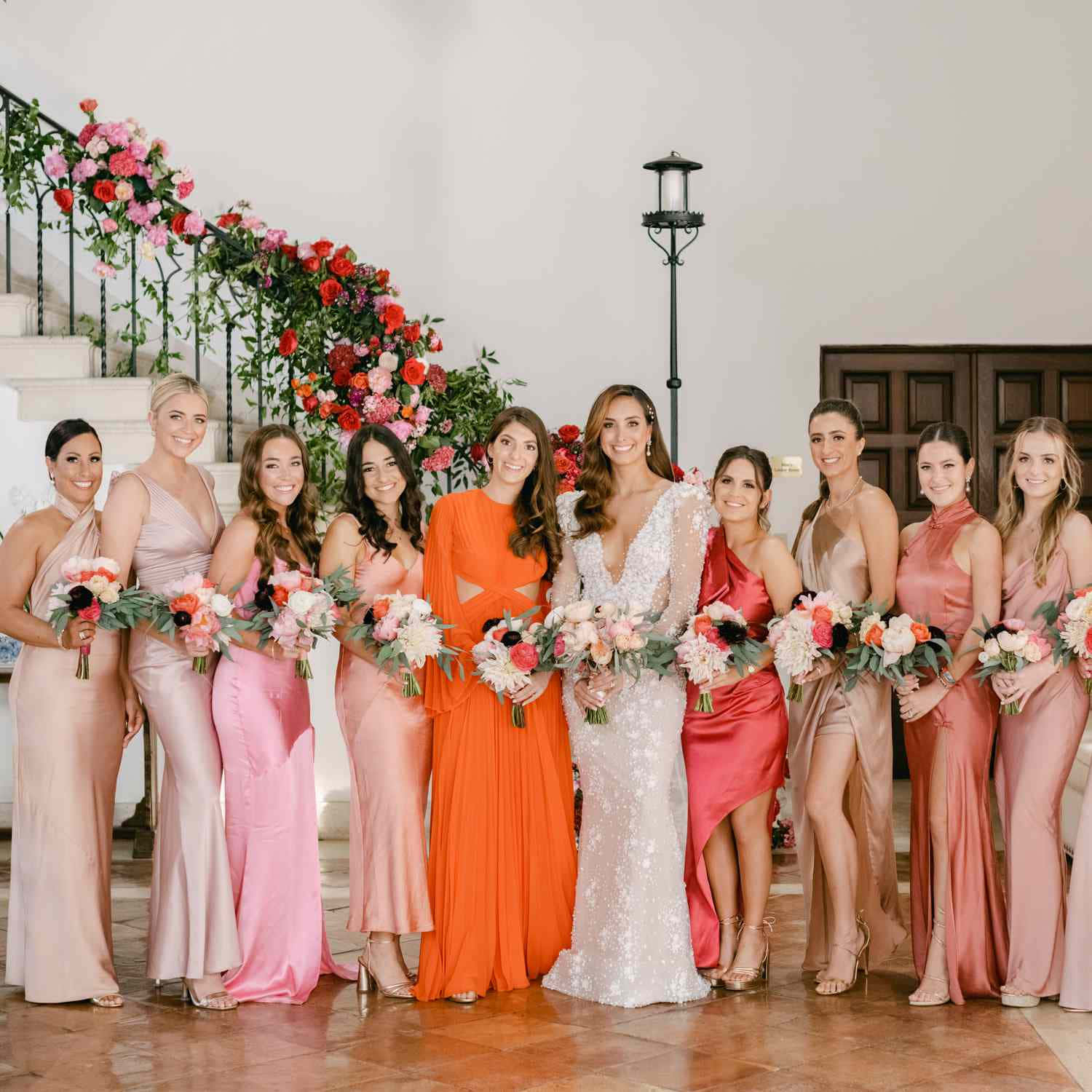 Bridal 桌布 壁紙 — 一位身穿閃亮白色婚紗的新娘，與穿著鮮豔粉紅色和橙色緞面禮服的伴娘們，手持花束，站在一個裝飾著紅色和粉紅色玫瑰瀑布的樓梯旁。