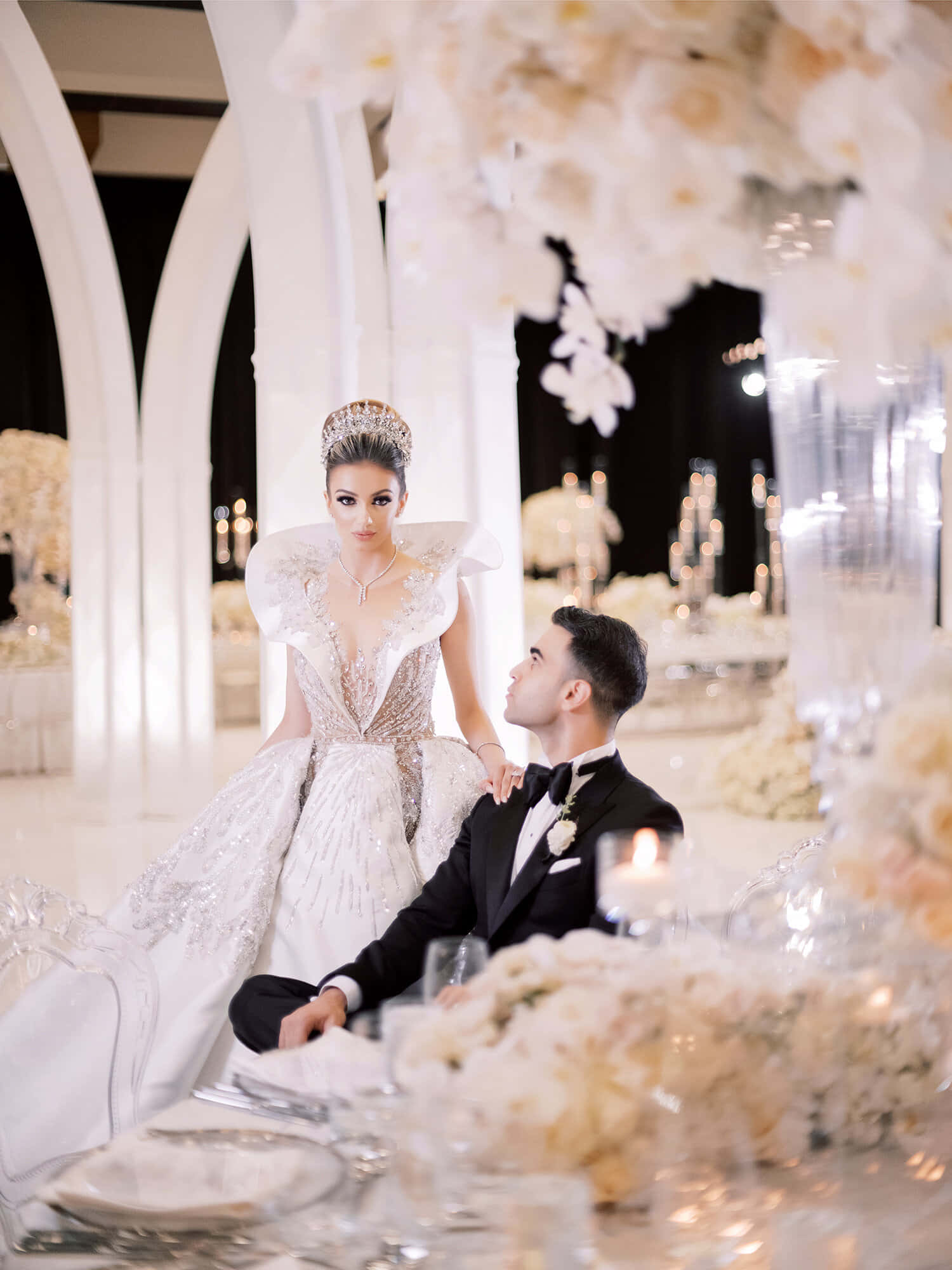 Black And White Wedding 桌布 壁紙 — 一位身穿閃亮白色婚紗的新娘站在一位身穿燕尾服、跪在地上的新郎上方，背景是華麗的白色花卉婚禮招待會。