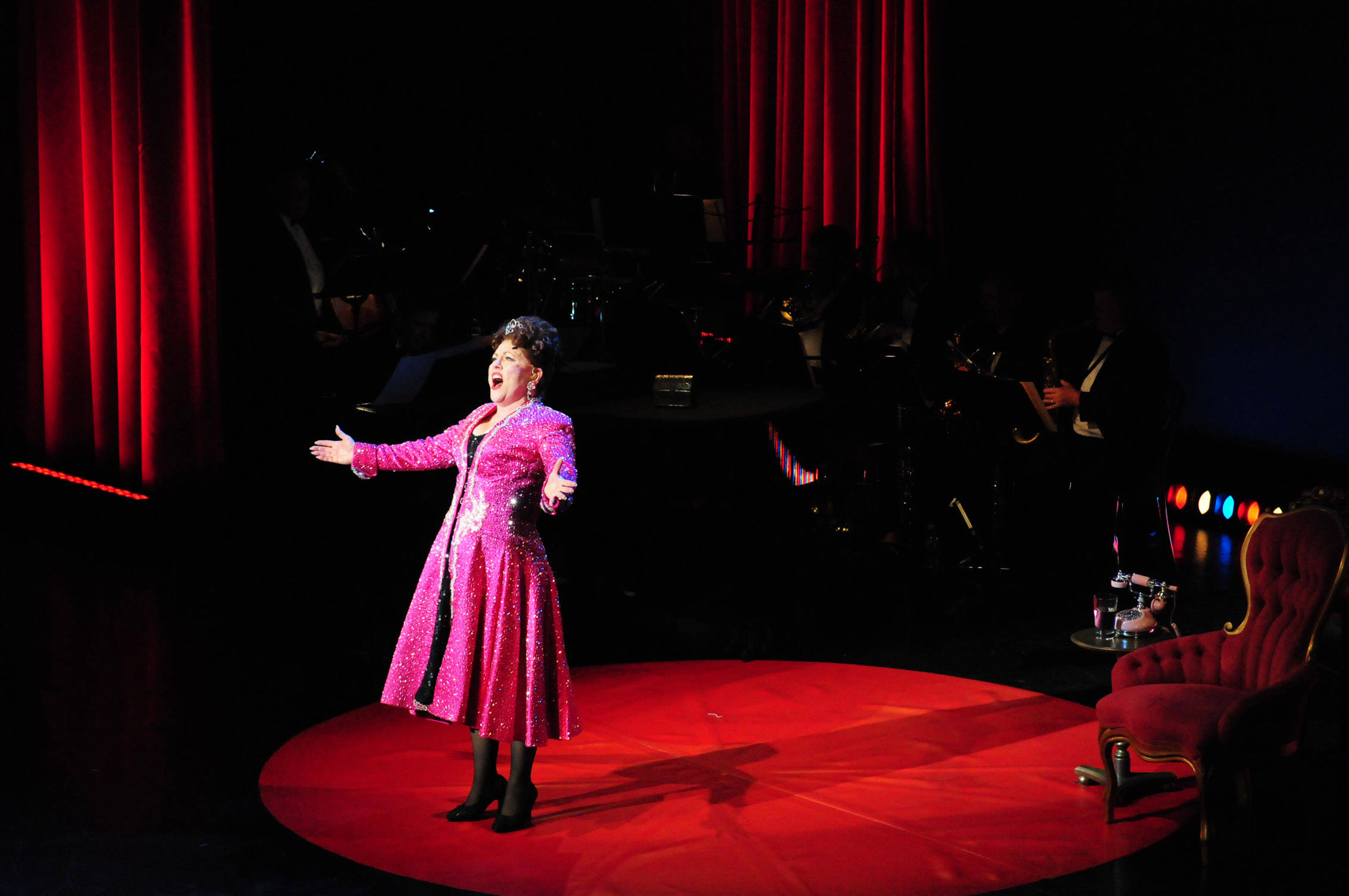 Singing 桌布 壁紙 — 一位身穿閃亮粉紅色外套的女性在紅色圓形舞台上，在戲劇性的聚光燈下，身後有樂隊，熱情地歌唱。