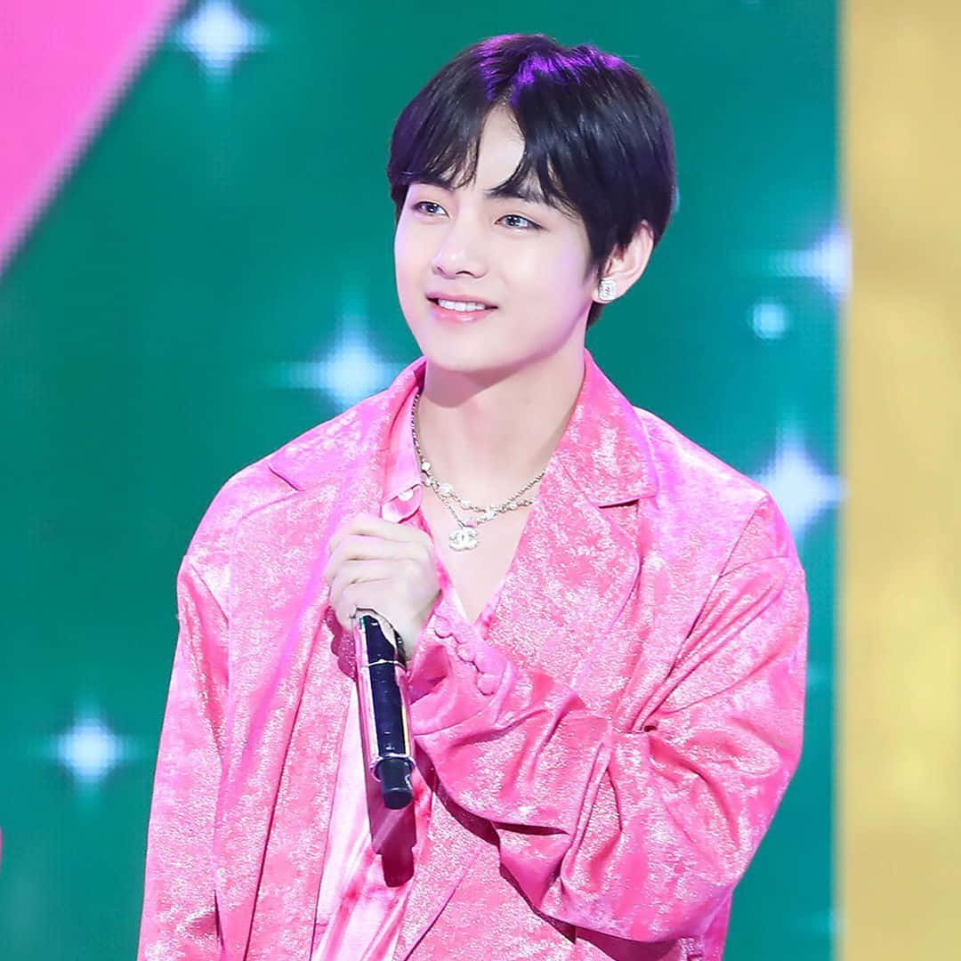 Taehyung Cute 桌布 壁紙 — 一位身穿閃亮粉紅色外套的年輕男子在色彩繽紛、星光閃爍的舞台上微笑著拿著麥克風。