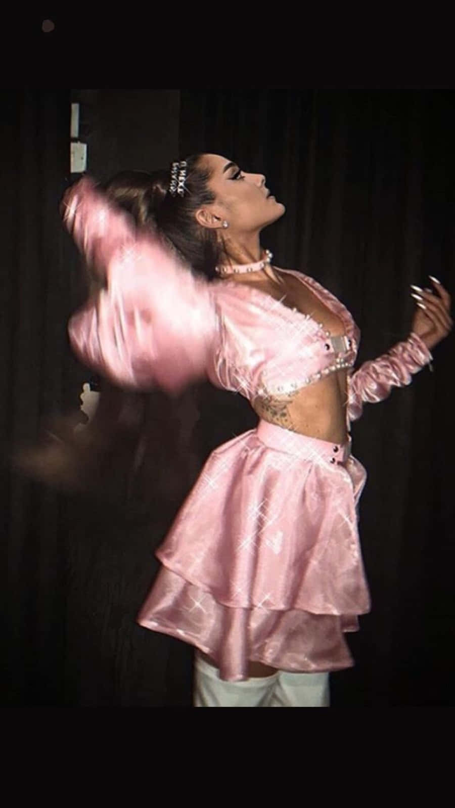 Aesthetic Ariana Grande 桌布 壁紙 — 一位身穿閃亮粉紅色服裝的女性在深色窗簾背景下旋轉，帶有動態模糊效果。