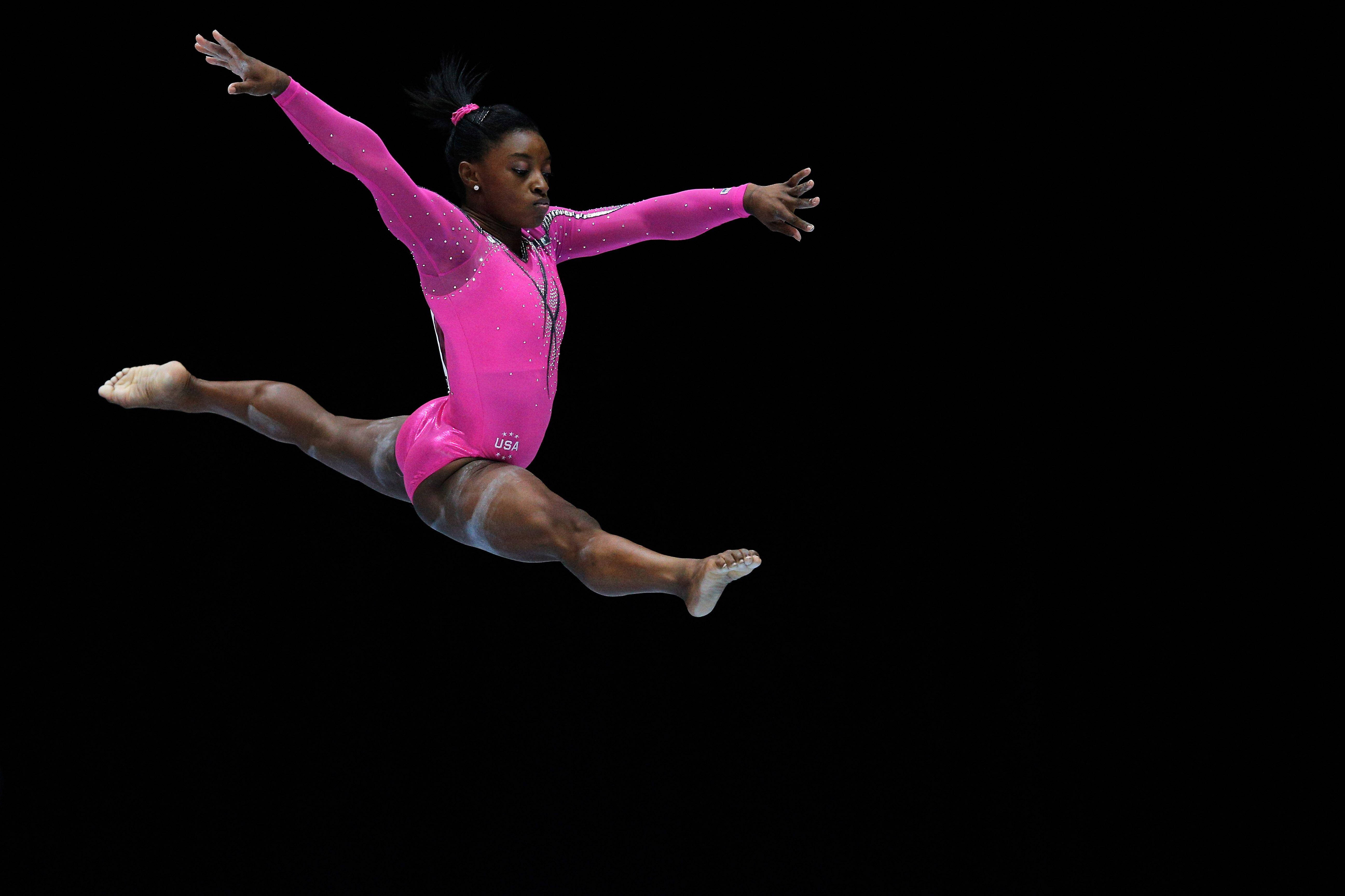 Simone Biles 桌布 壁紙 — 一位身穿閃亮粉紅色緊身衣的女體操運動員在純黑色背景上執行動態劈叉跳躍。