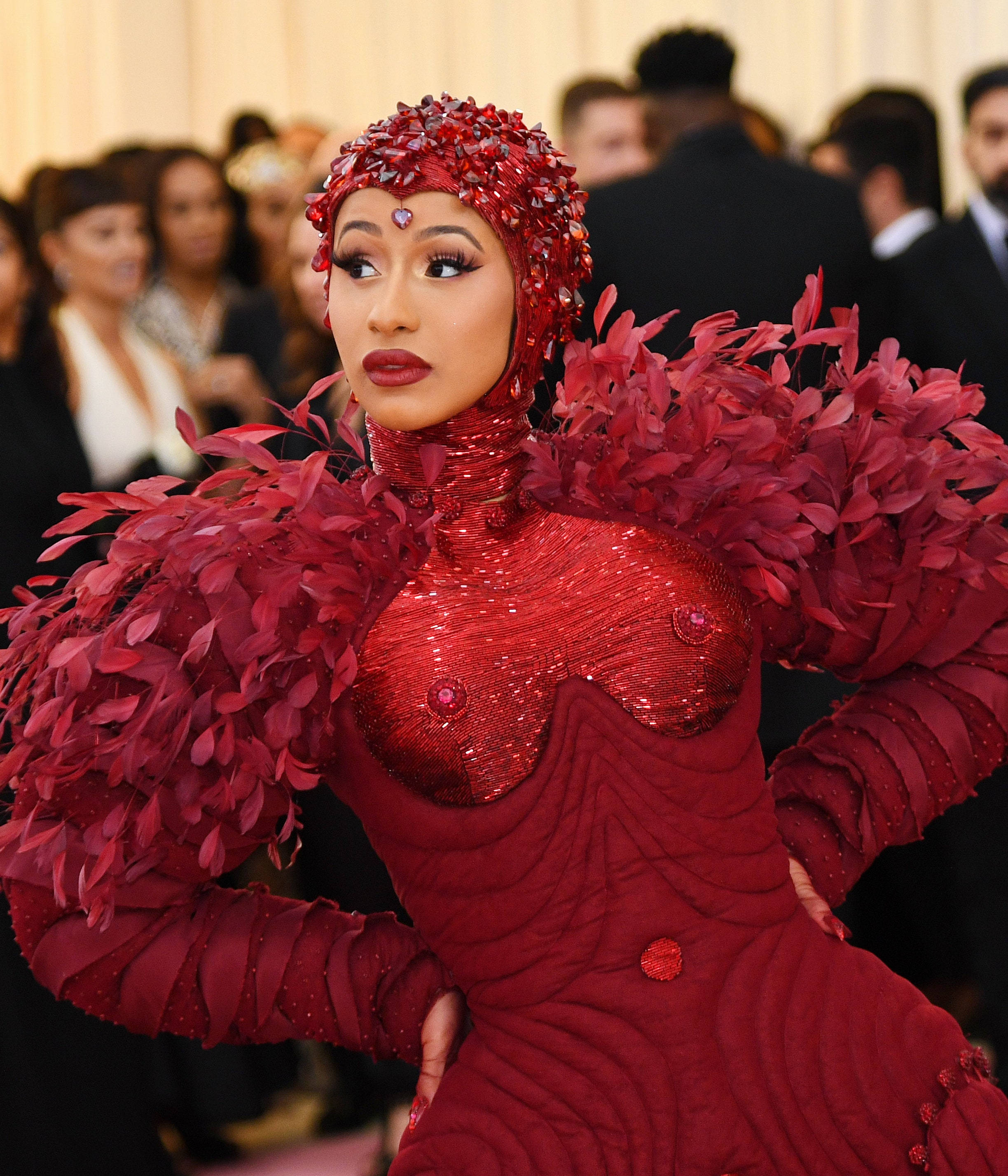 cardi 桌布 壁紙 — 一位身穿閃亮紅色亮片禮服、搭配羽毛袖子和相配頭飾的女性，在華麗的活動中自信地擺姿勢。