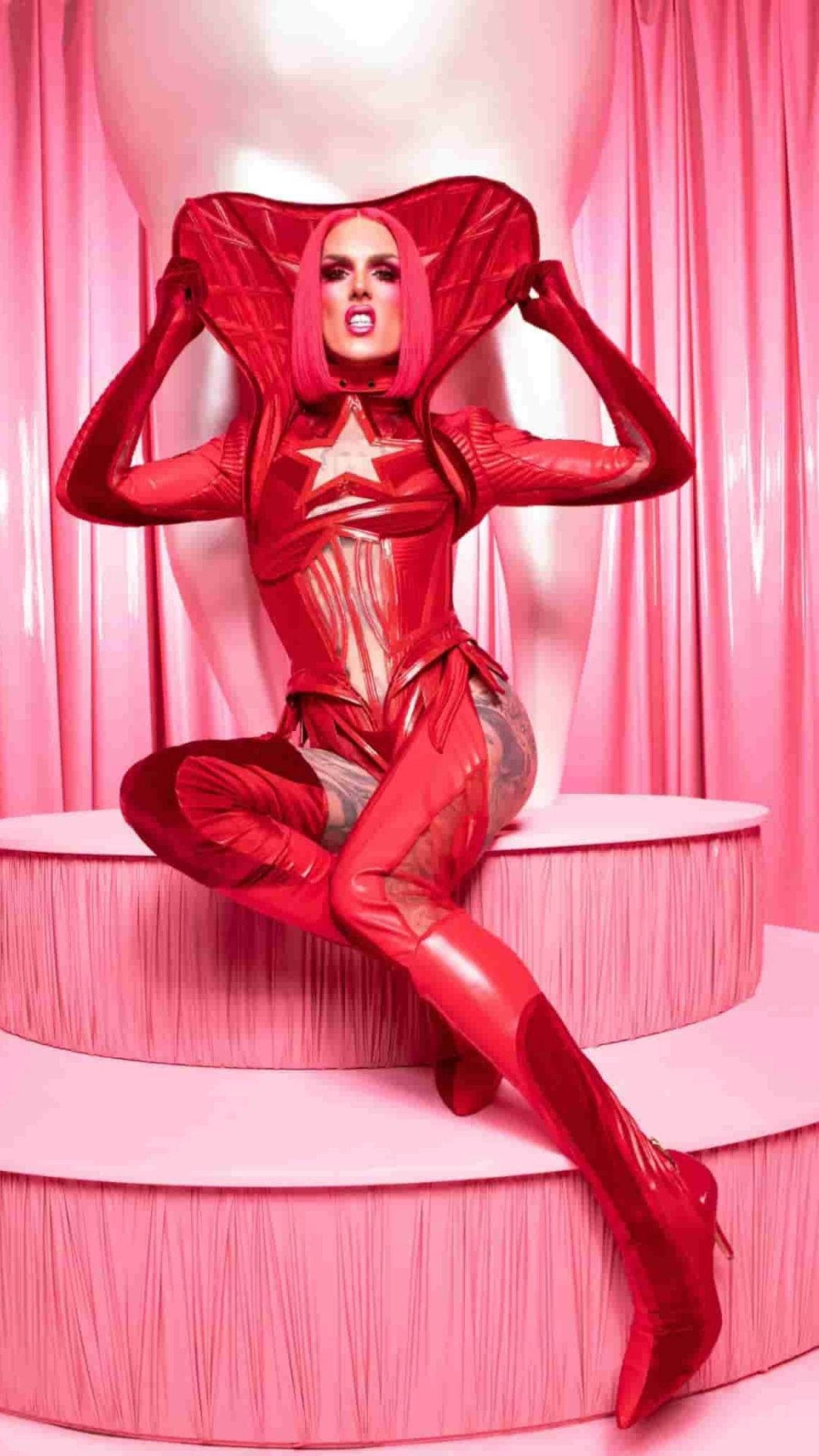Jeffree Star 桌布 壁紙 — 一位身穿閃亮紅色超級英雄服裝、帶有星星標誌的女性，自信地坐在粉紅色階梯上，背景是粉紅色窗簾。