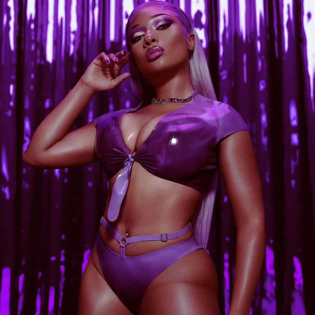 Megan Thee Stallion 桌布 壁紙 — 一位身穿閃亮紫色服裝的女性在滴落的紫色窗簾背景前自信地擺姿勢。