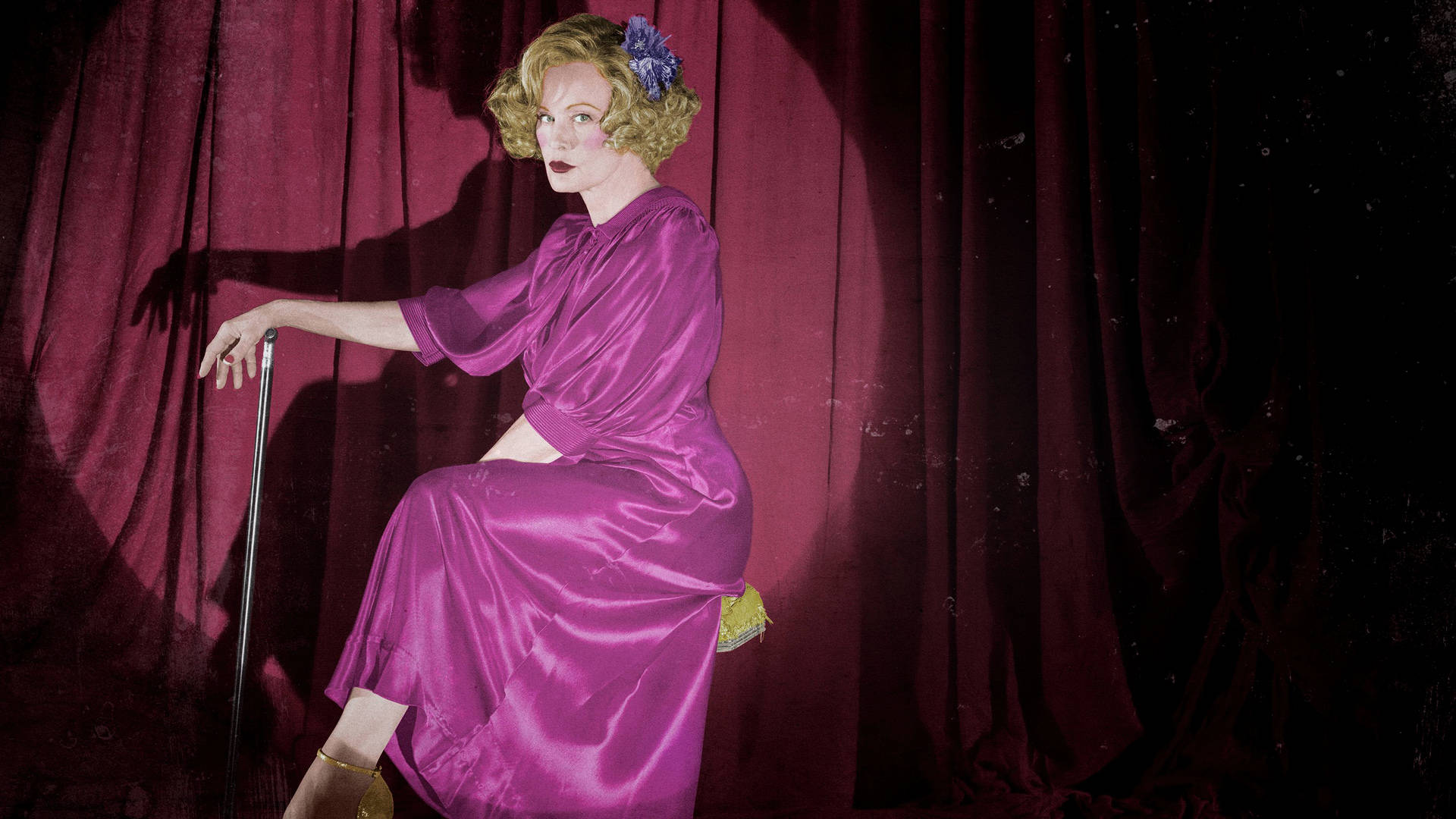 American Horror Story 桌布 壁紙 — 一位身穿閃亮紫色緞面長裙的女性，在戲劇性的舞台燈光下，優雅地坐在手杖上，背景是深紅色天鵝絨窗簾。