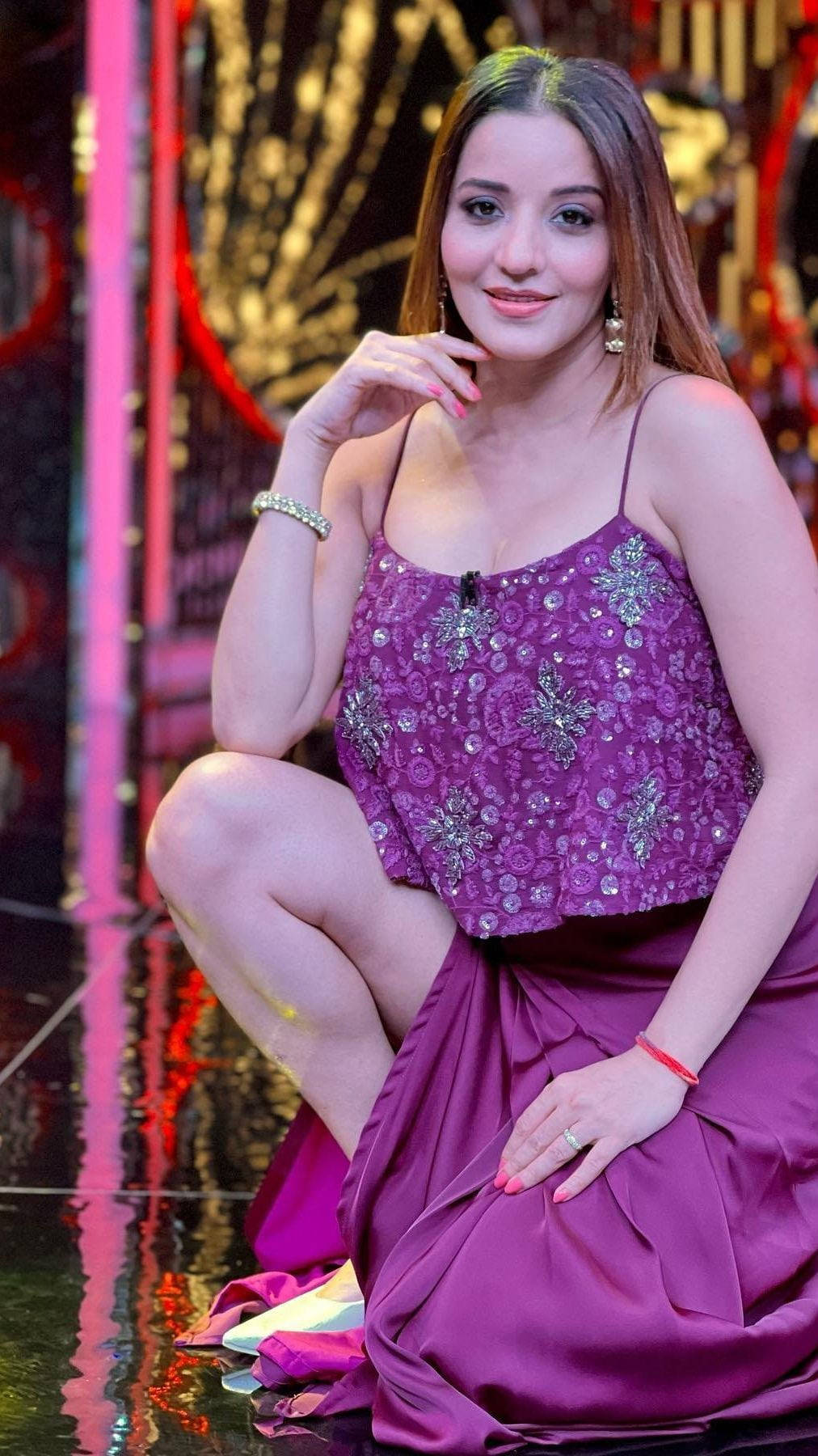Bhojpuri 桌布 壁紙 — 一位身穿閃亮紫色連衣裙的女性在反射性舞台上優雅地擺姿勢，身後有充滿活力的粉紅色和金色燈光。