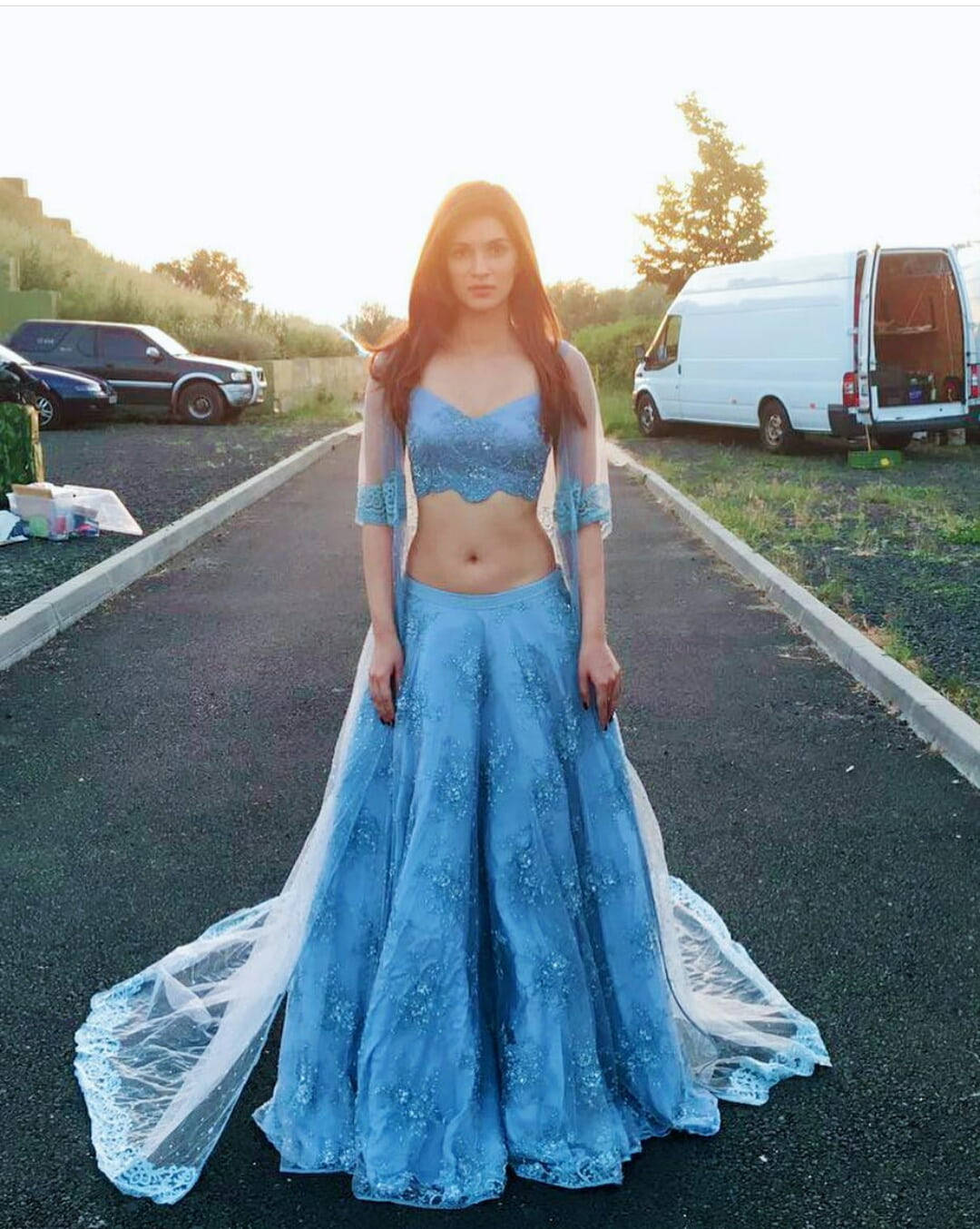 洋裝 桌布 壁紙 — 一位身穿閃亮藍色 lehenga 的女性站在瀝青路上，正值黃金時段，附近停有車輛。