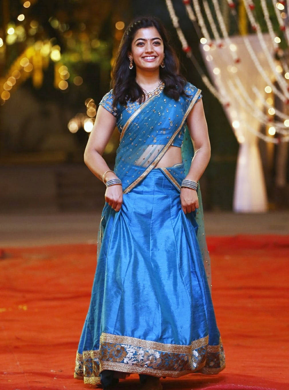 telugu 桌布 壁紙 — 一位身穿閃亮藍色刺繡金色 lehenga 的燦爛女性，在節日燈串下的紅地毯上燦爛微笑。