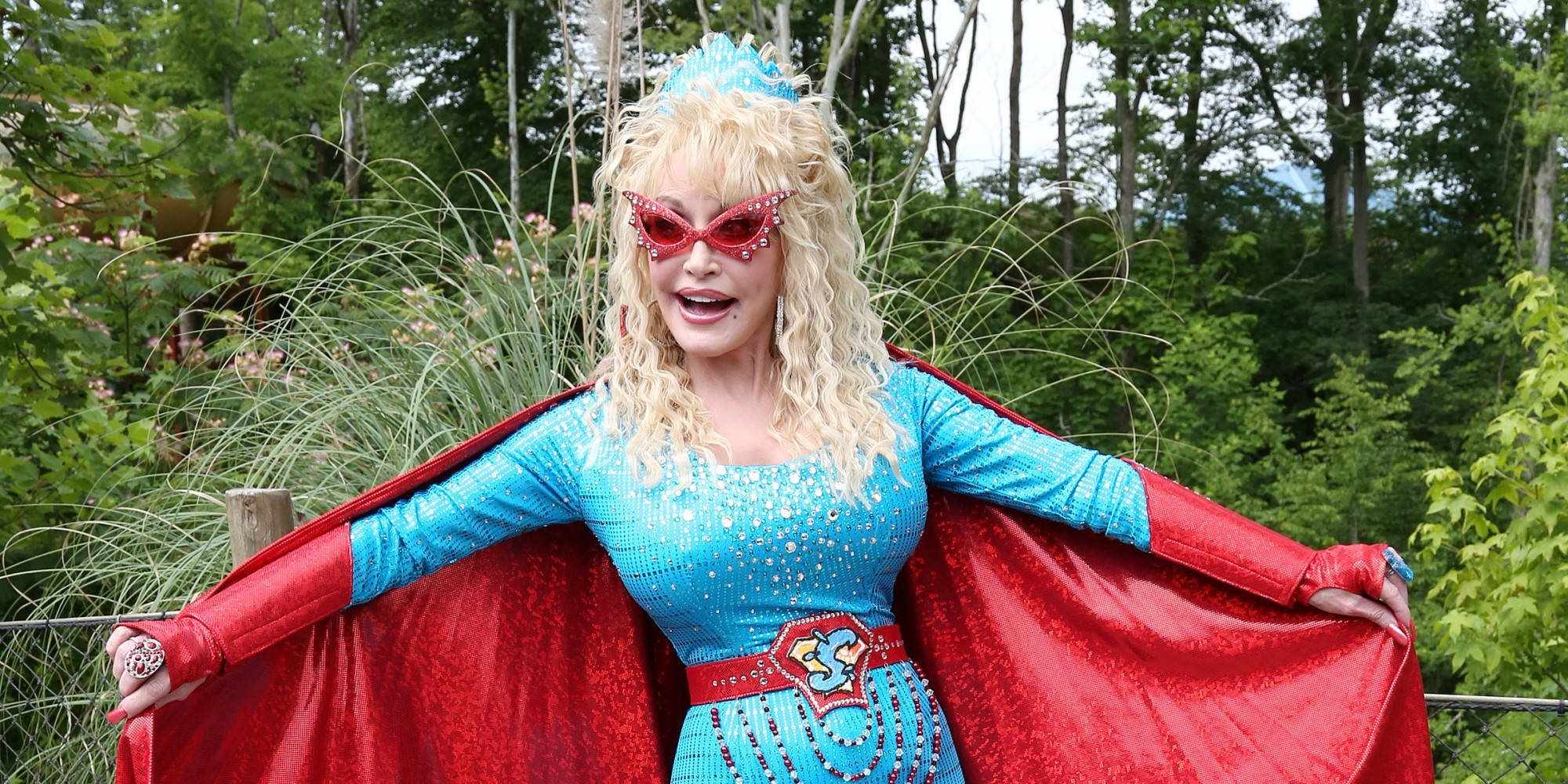 dolly 桌布 壁紙 — 一位身穿閃亮藍色超級英雄服裝、披著紅色斗篷、戴著粉紅色太陽眼鏡的女性，愉快地站在茂密的綠色森林中。