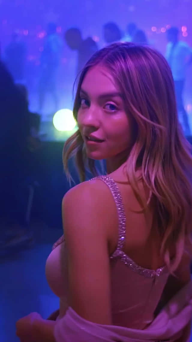 Euphoria Hbo iPhone 桌布 壁紙 — 一位身穿閃亮連衣裙的女性在霓虹燈閃爍的夜店中轉身回頭看，身後有模糊的舞者。