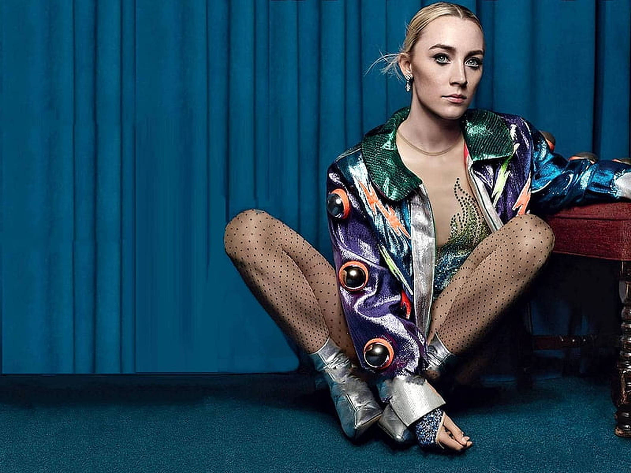 Saoirse Ronan 桌布 壁紙 — 一位身穿閃亮金屬外套和波點緊身褲的女性，端坐在深藍色窗簾背景前。