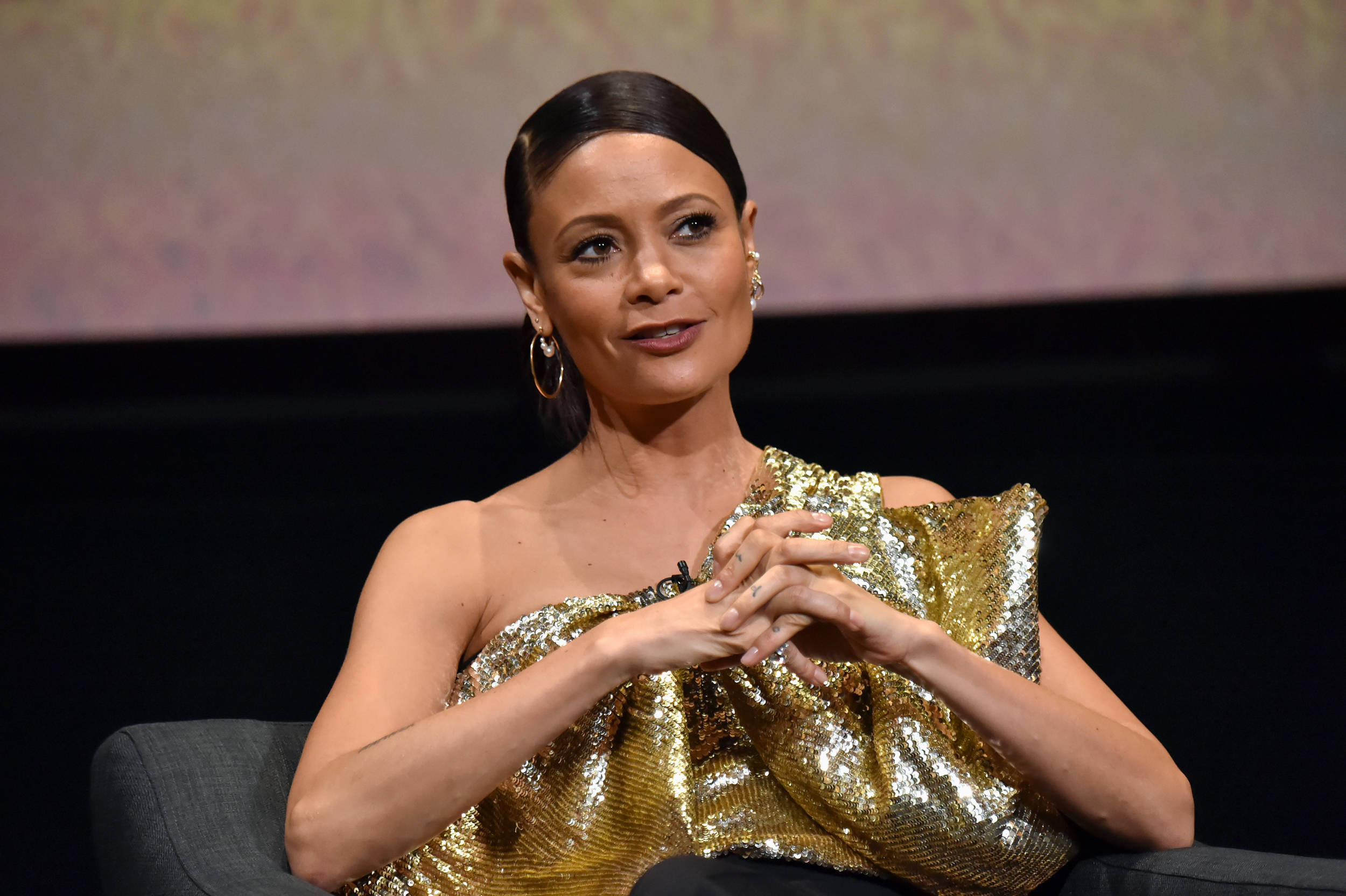 Actress HD 桌布 壁紙 — 一位身穿閃亮金色單肩連衣裙的女性，在柔和漸變背景的活動中，優雅地坐在深色椅子上，雙手交握。