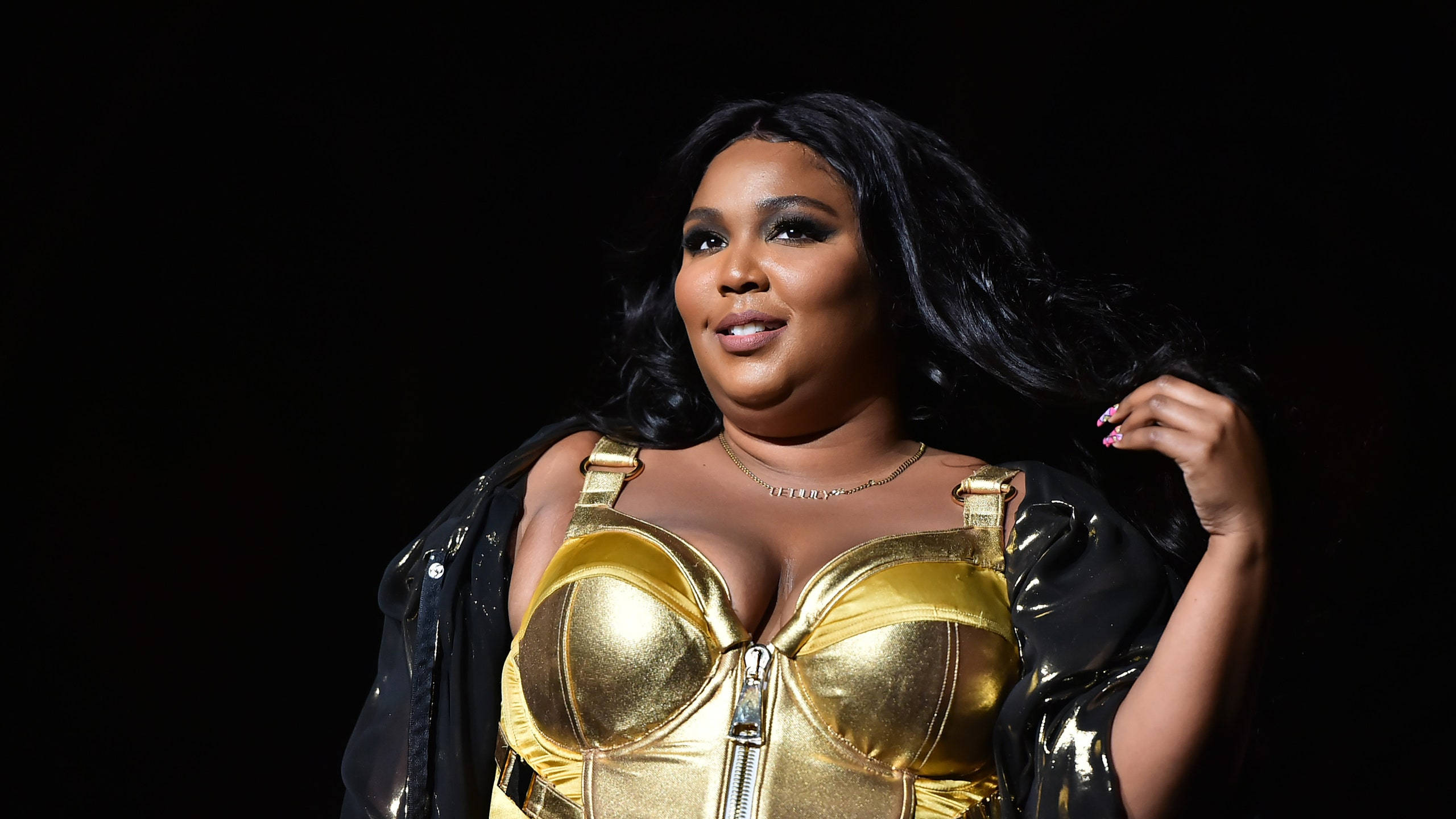 Lizzo 桌布 壁紙 — 一位身穿閃亮金色緊身胸衣和黑色外套的女性，在深色背景下自信地擺姿勢，頭髮飄逸。