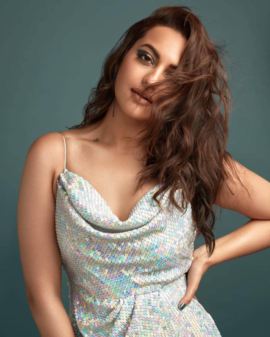 Sonakshi Sinha 桌布 壁紙 — 一位身穿閃亮銀色亮片連衣裙的女性，在柔和的青綠色工作室背景前，自信地擺出姿勢，頭髮隨風飄揚。