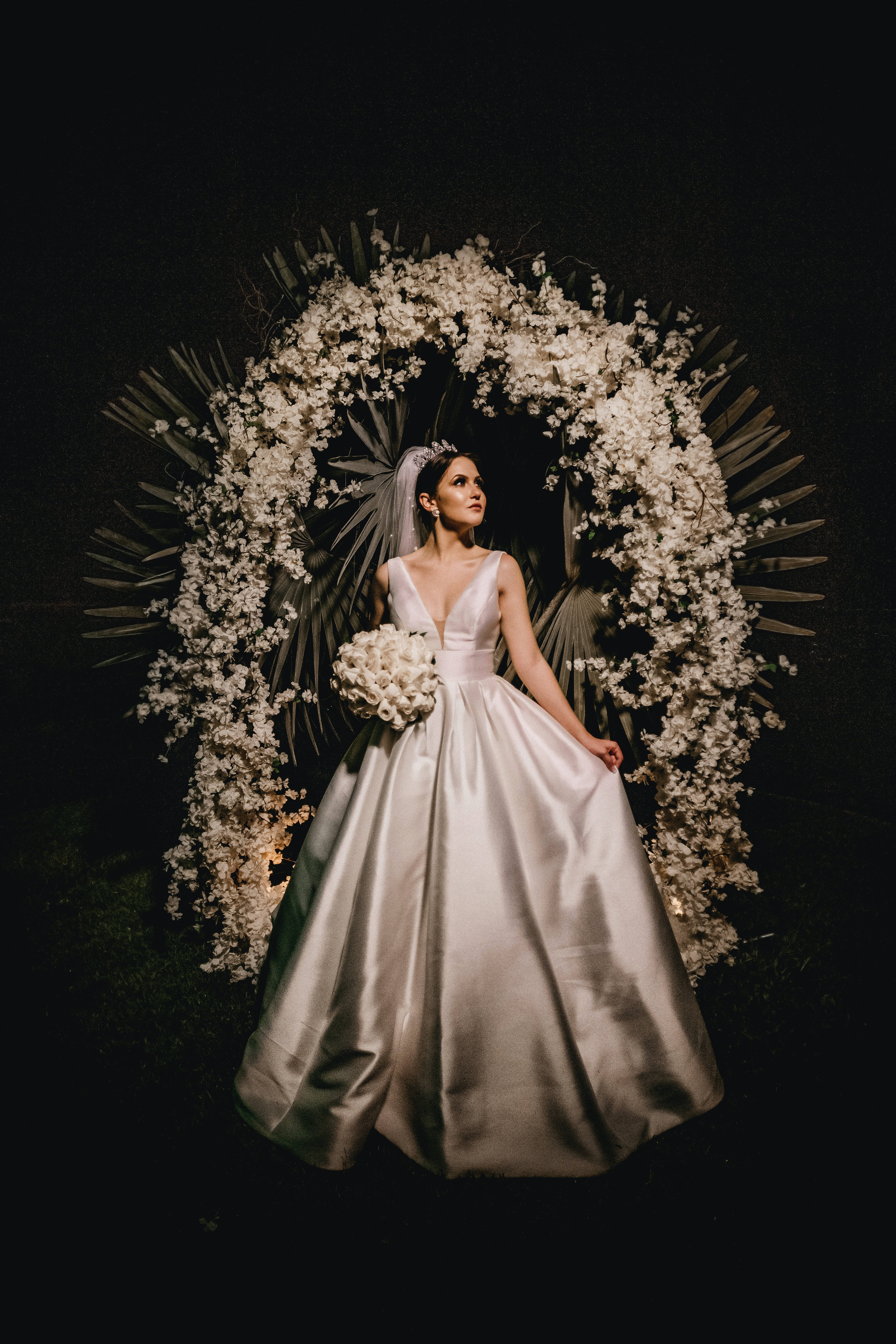 Bridal 桌布 壁紙 — 一位身穿閃亮銀色禮服的新娘，在夜晚優雅地站在璀璨的花拱門下，手持一束白色鮮花。