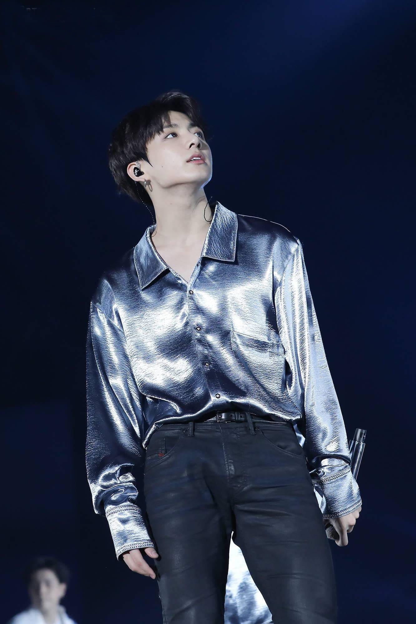 BTS 桌布 壁紙 — 一位身穿閃亮銀色襯衫和黑色褲子的男性表演者站在黑暗的舞台上，手持麥克風向上看。