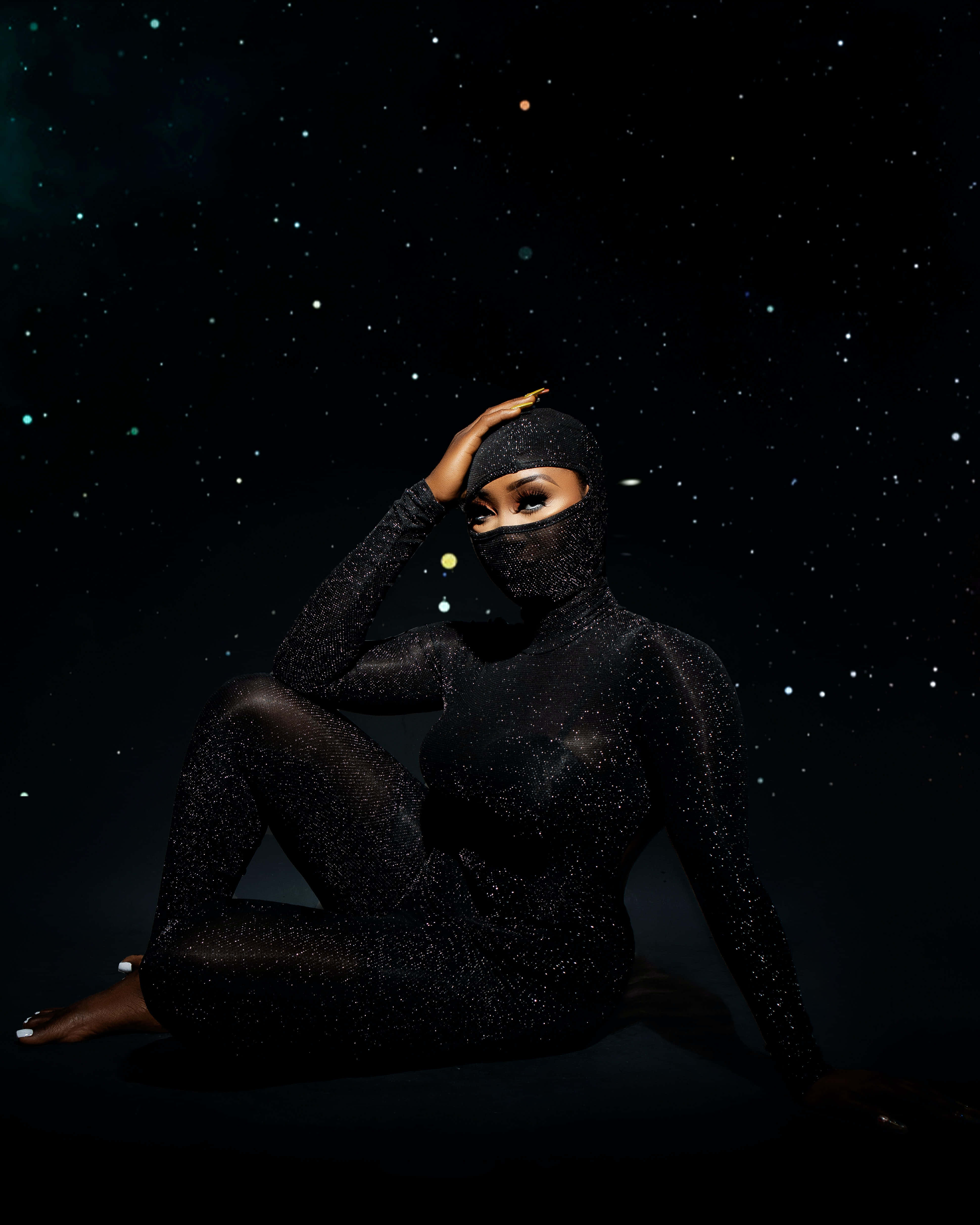 黑人女性 桌布 壁紙 — 一位身穿閃亮黑色連體衣的女性，在星空宇宙背景下優雅地坐著，一手放在頭上。