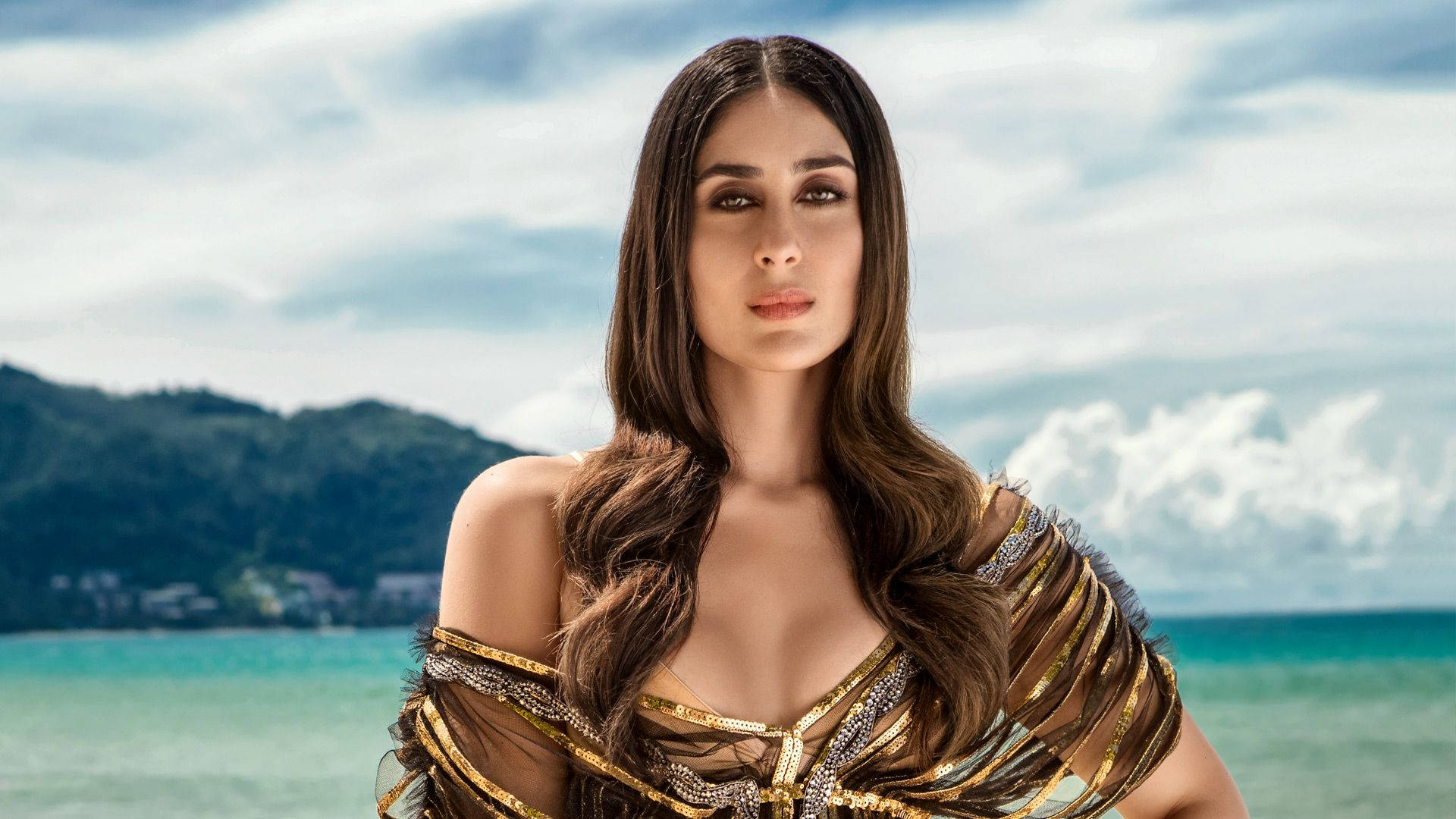 Kareena 桌布 壁紙 — 一位身穿閃爍金銀服裝的女性，自信地站在藍綠色海洋和茂密綠色山丘的背景前，天空多雲。
