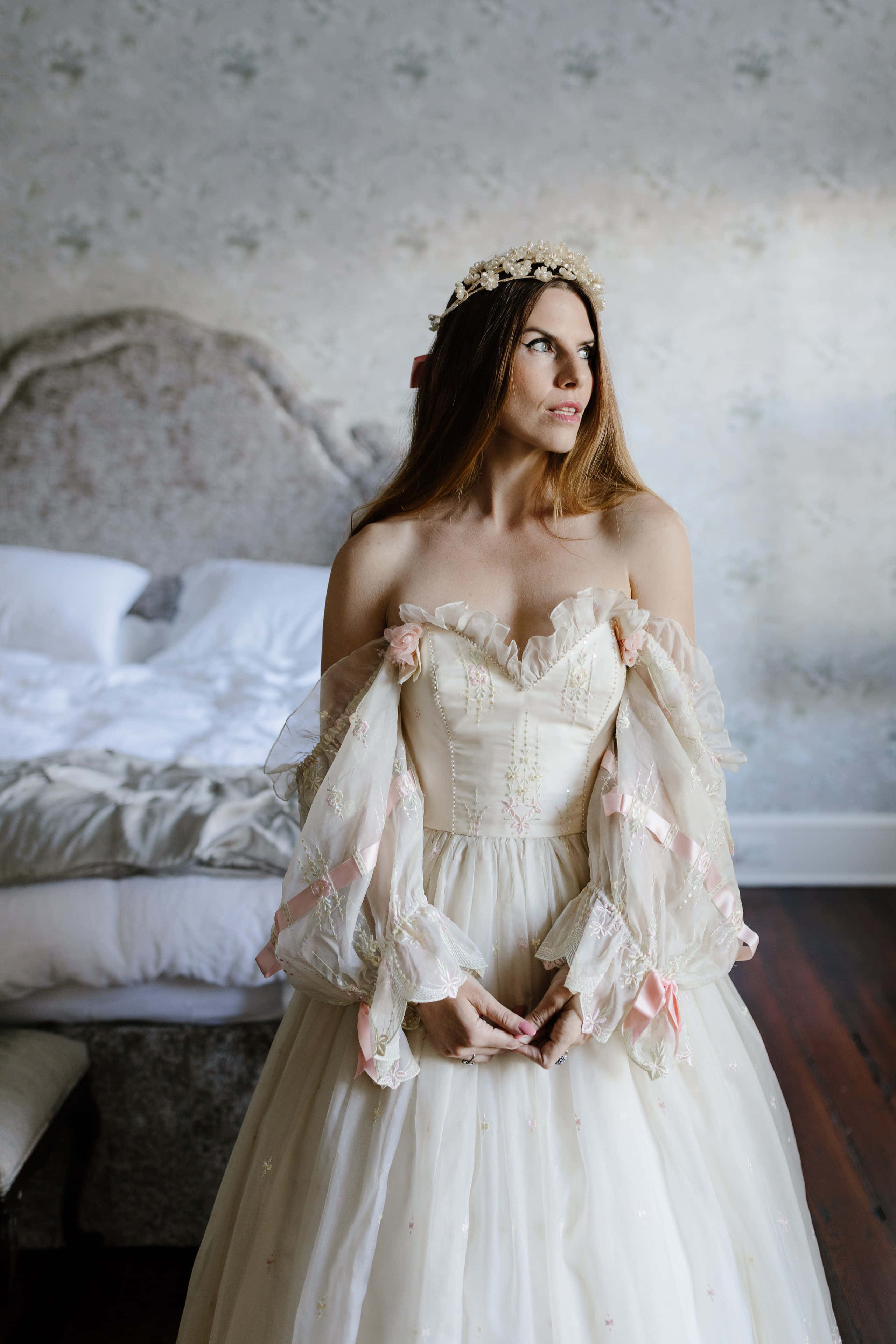 Bride's Dress 桌布 壁紙 — 一位身穿露肩象牙色禮服、繫著粉紅色絲帶的女性，站在燈光柔和、鋪有花卉壁紙和復古床的臥室中。