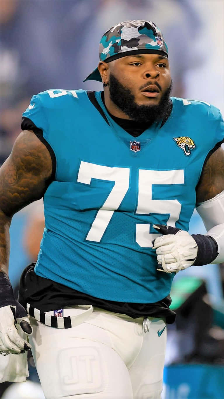 Jawaan Taylor 桌布 壁紙 — 一位身穿青綠色75號球衣的傑克遜維爾美洲虎足球員在明亮的燈光下於體育場奔跑。