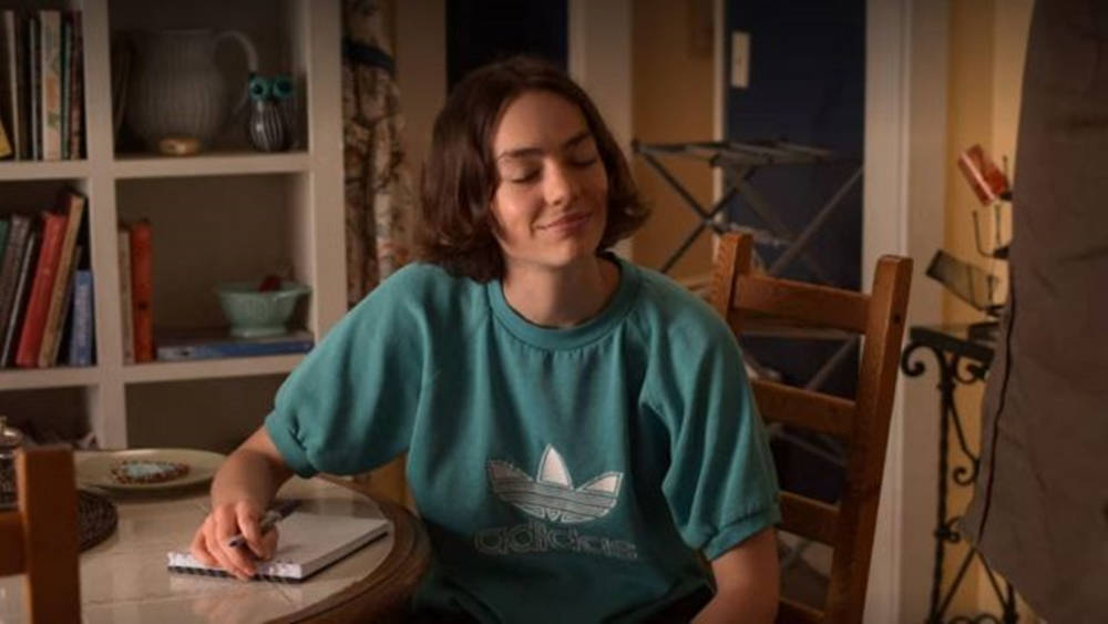 Netflix 桌布 壁紙 — 一位身穿青綠色Adidas運動衫的年輕女性，在溫暖燈光的家中木桌上寫筆記本，平靜地微笑。