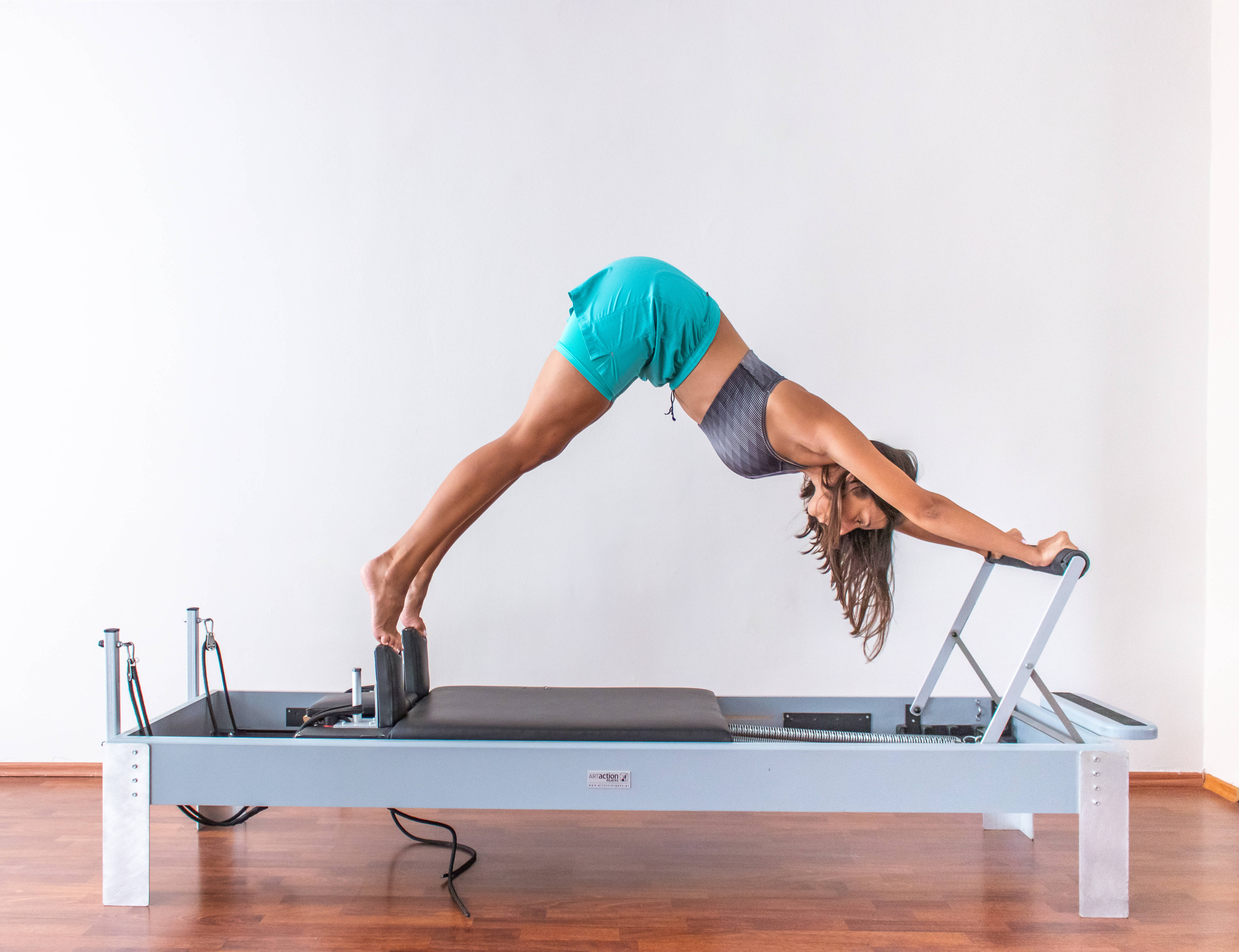 Exercise 桌布 壁紙 — 一位身穿青綠色短褲和灰色上衣的女性，在簡約的白色牆壁前做皮拉提斯核心床橋式動作。