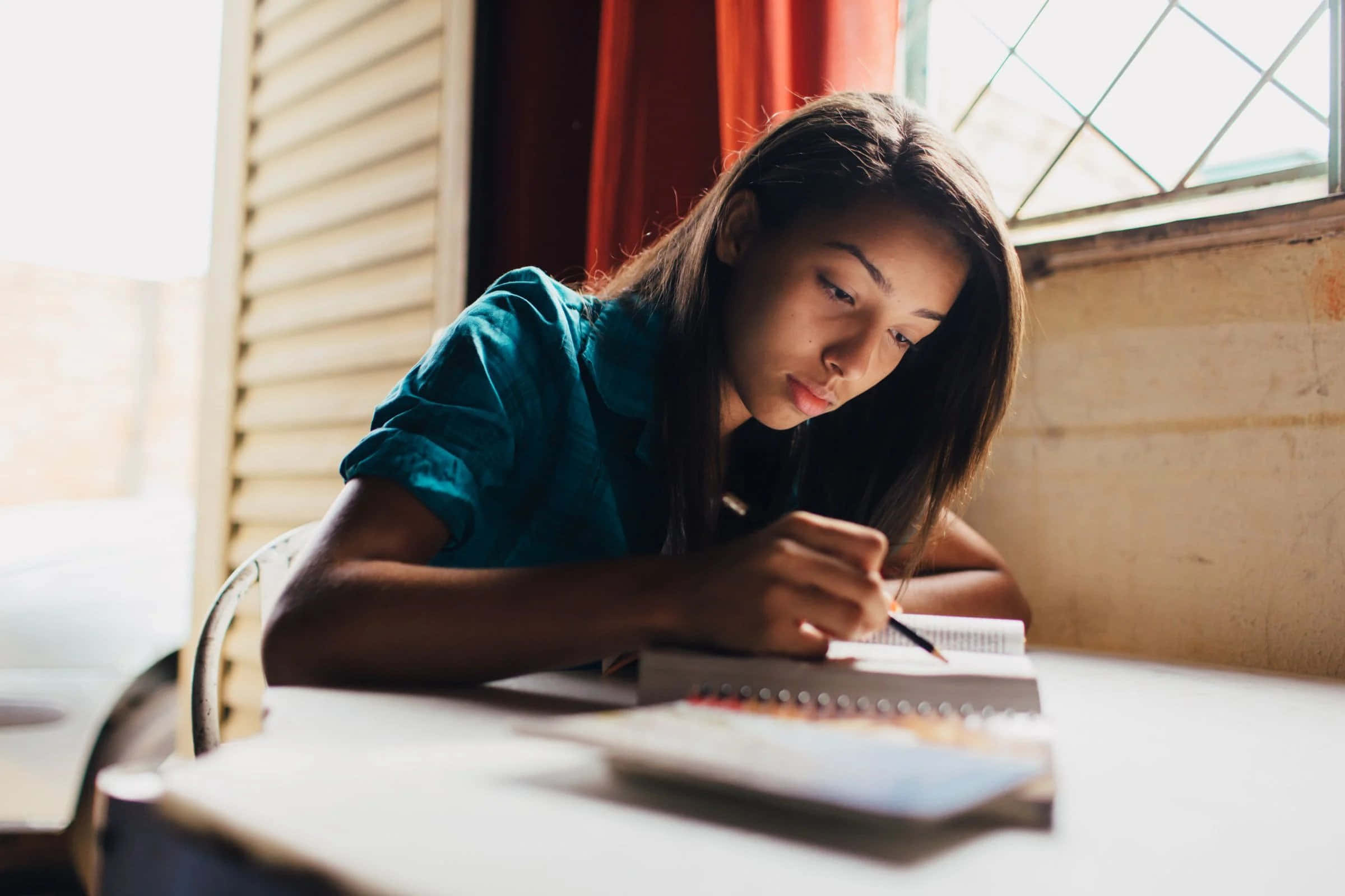 Study Aesthetic 桌布 壁紙 — 一位身穿青綠色襯衫的年輕女子，在陽光照射的木桌旁、靠近掛有紅色窗簾的窗戶，專注地寫筆記。