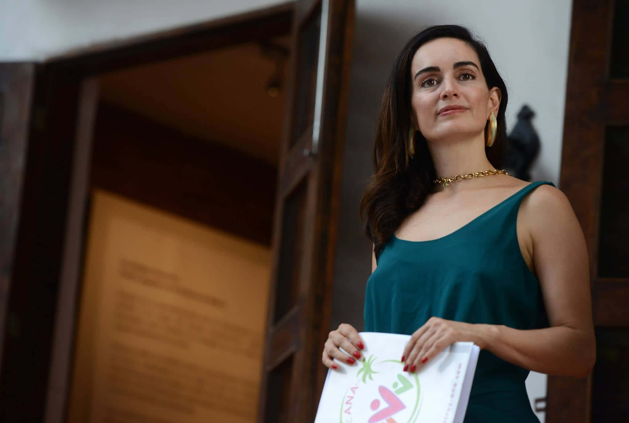 Ana De La Reguera 桌布 壁紙 — 一位身穿青綠色連衣裙的優雅女士，站在溫暖燈光的木門前，手持一本品牌小冊子。