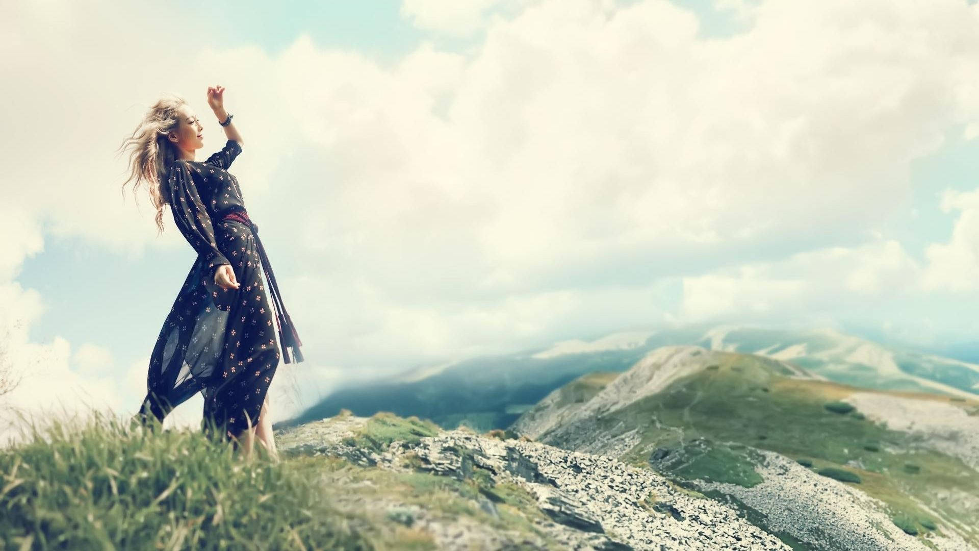 태연 桌布 壁紙 — 一位身穿飄逸深色連衣裙的女性赤腳站在長滿青草的山脊上，向明亮的雲天舉起手。