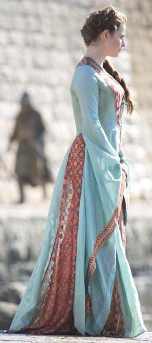 Esmé Bianco 桌布 壁紙 — 一位身穿飄逸淺藍色和紅色圖案長袍的女性，側身站在模糊的石牆背景前。