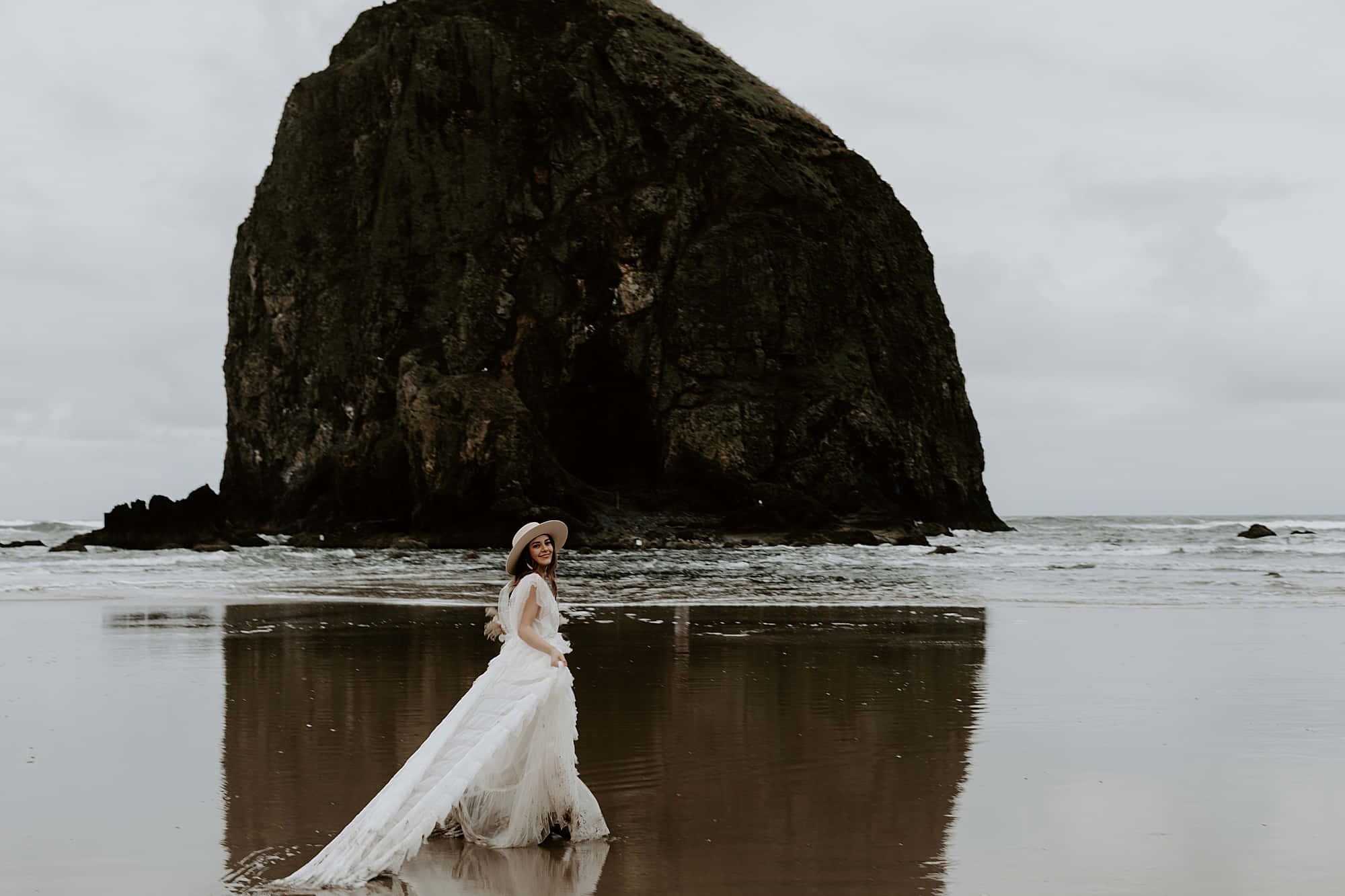 Oregon Coast 桌布 壁紙 — 一位身穿飄逸白色婚紗和寬邊帽的女子在潮濕的海灘上行走，身後有一座巨大的黑色岩石聳立，天空多雲。