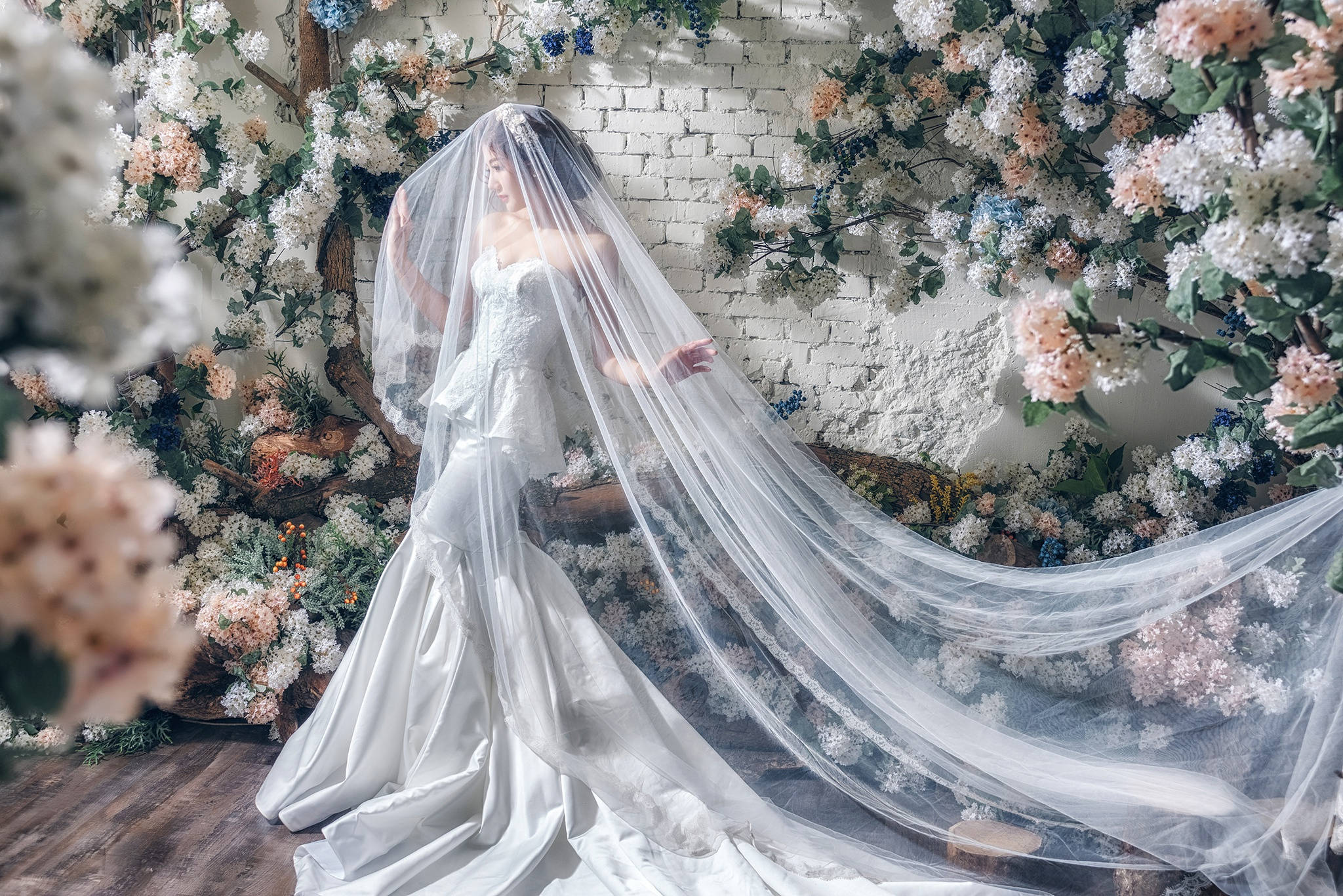 Wedding 桌布 壁紙 — 一位身穿飄逸白色婚紗和頭紗的新娘，在白色磚牆前，優雅地站在茂盛的花拱門中。