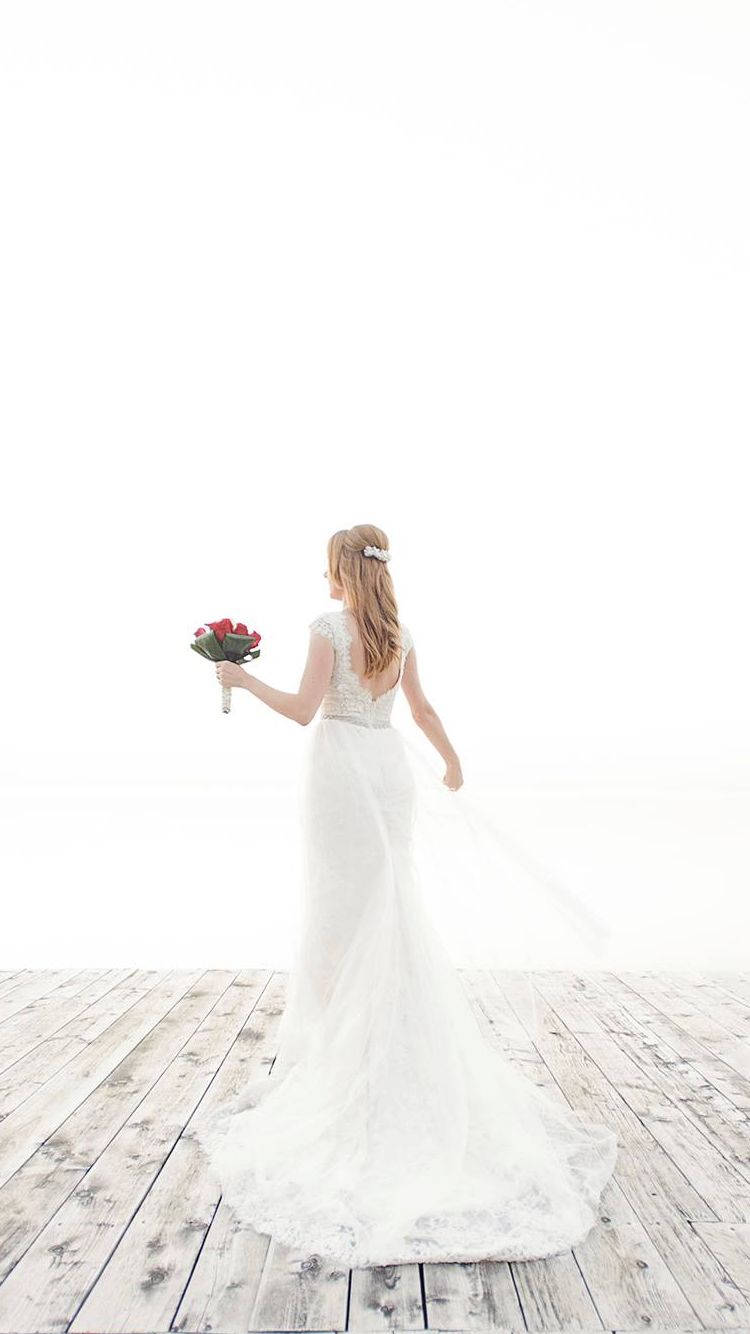 Wedding Dress 桌布 壁紙 — 一位身穿飄逸白色婚紗的新娘，在風化的木甲板上手持一束紅玫瑰，背對著明亮的白色背景走開。
