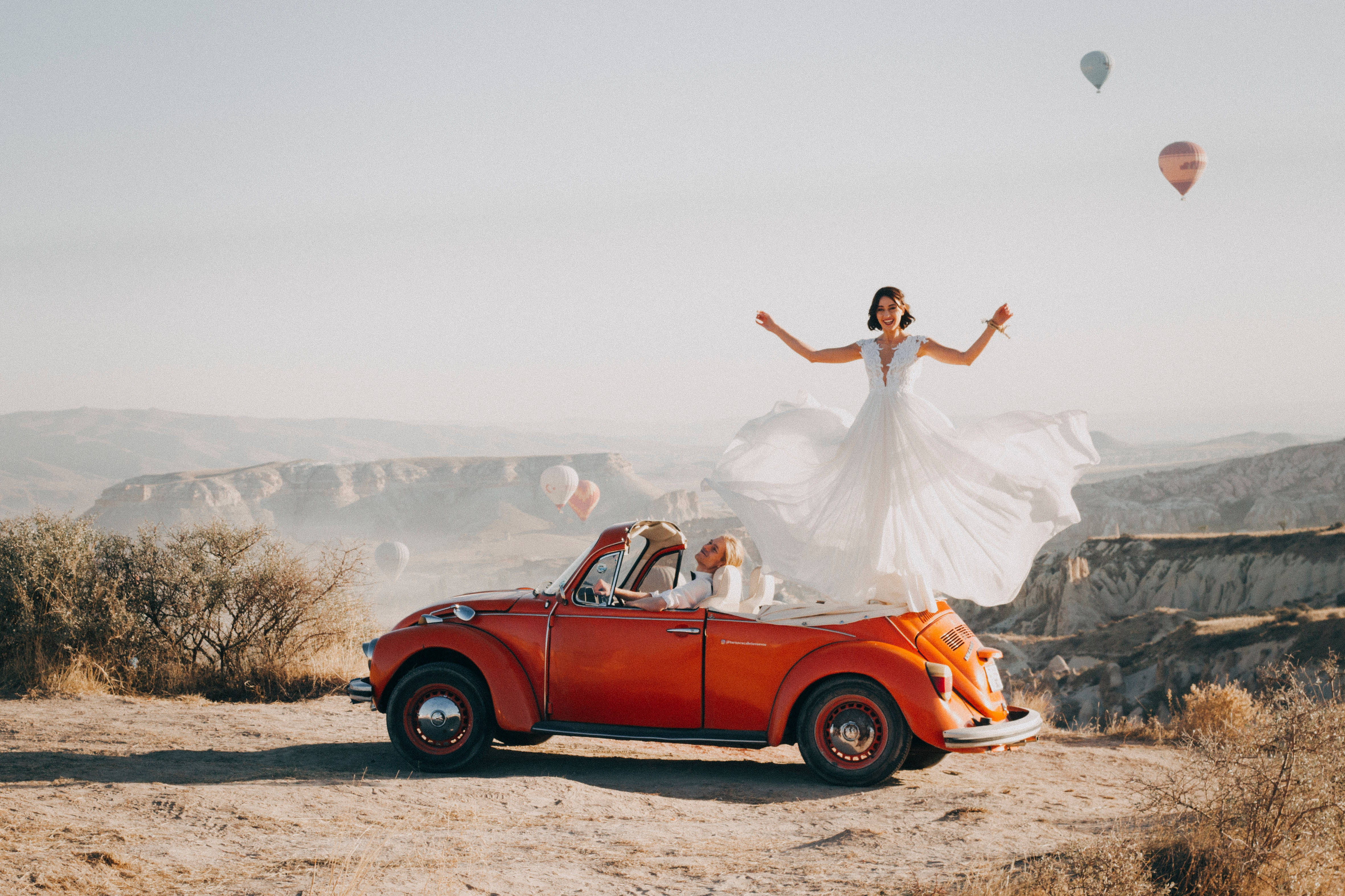 Bridal 桌布 壁紙 — 一位身穿飄逸白色婚紗的新娘，快樂地跳躍在一輛復古紅色敞篷車上方，車內有新郎，熱氣球在崎嶇的峽谷景觀上空飄浮。