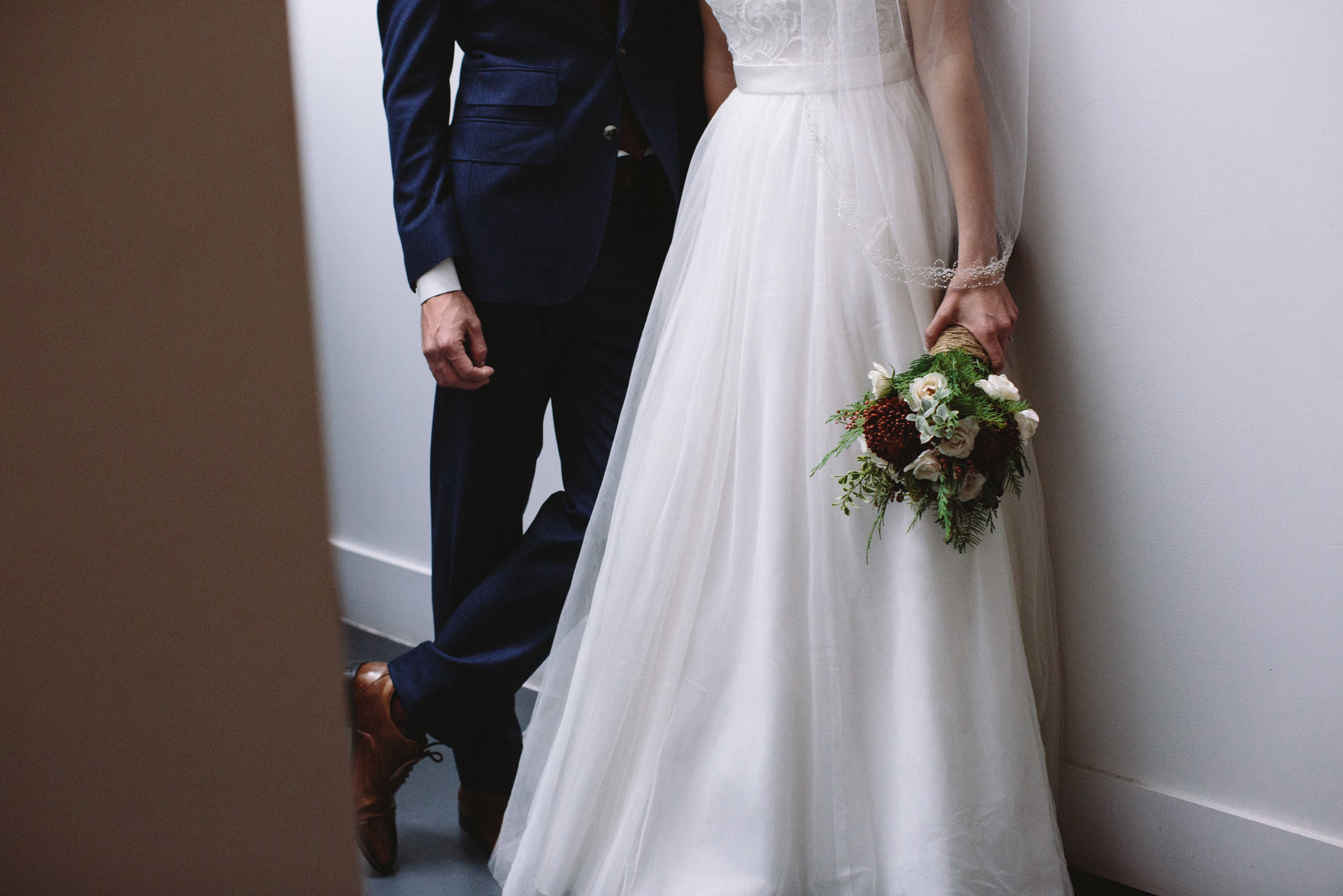 Groom 桌布 壁紙 — 一位身穿飄逸白色婚紗的新娘，手持豐盛的花束，站在身穿海軍藍西裝的新郎旁邊，背景是素牆。