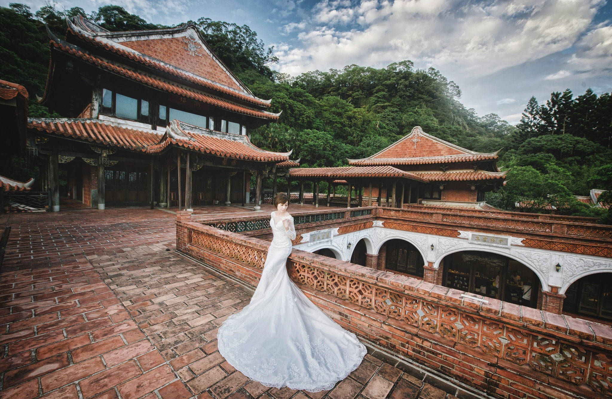 Wedding Dress 桌布 壁紙 — 一位身穿飄逸白色婚紗的新娘，站在鋪有赤陶磚的陽台上，俯瞰著傳統東亞寺廟建築，這些建築坐落在鬱鬱蔥蔥的綠色山丘中，天空部分多雲。