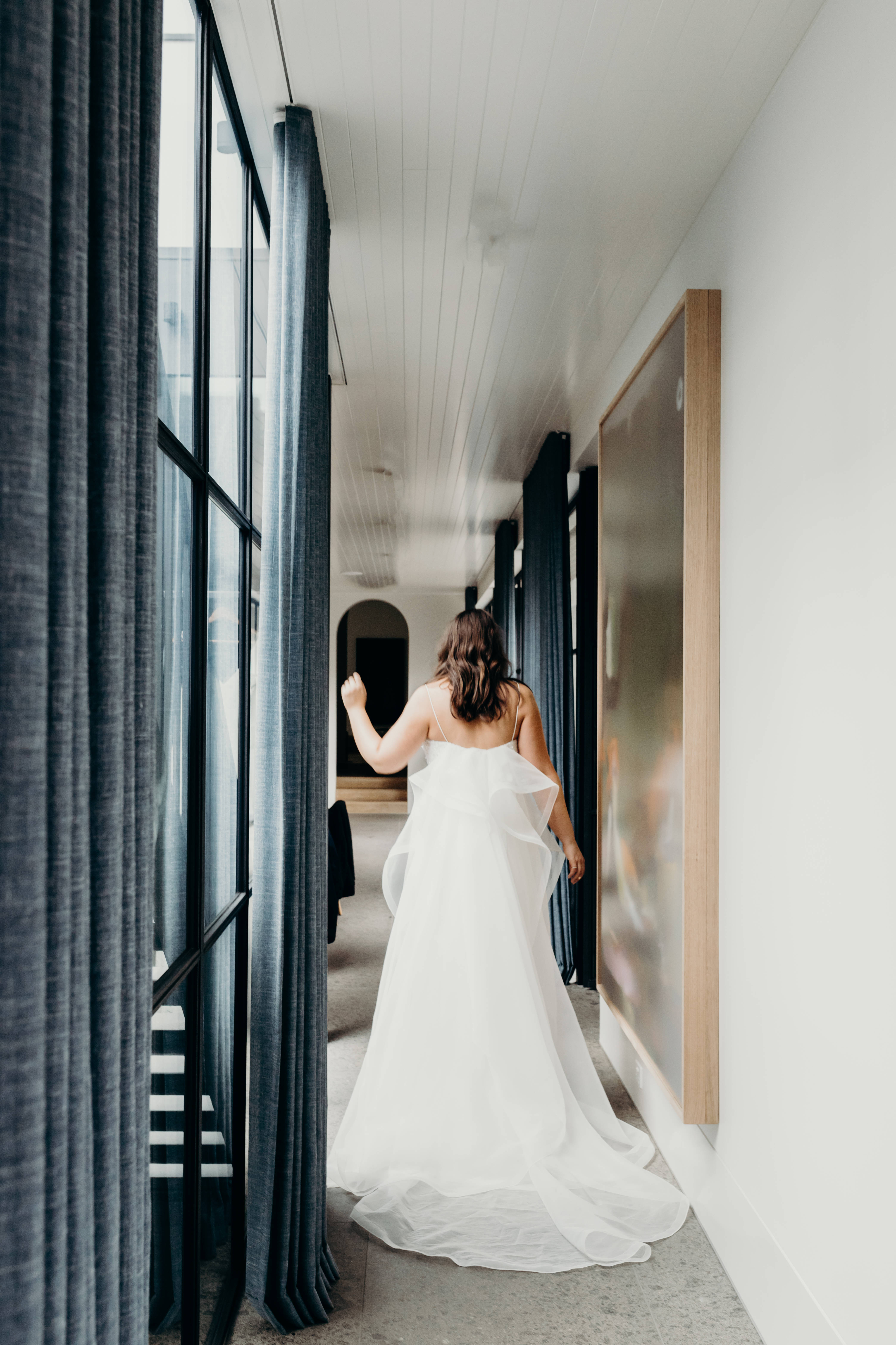 Wedding Aesthetic 桌布 壁紙 — 一位身穿飄逸白色婚紗的新娘，走在一條現代化的走廊上，兩旁是大窗戶和深色窗簾。