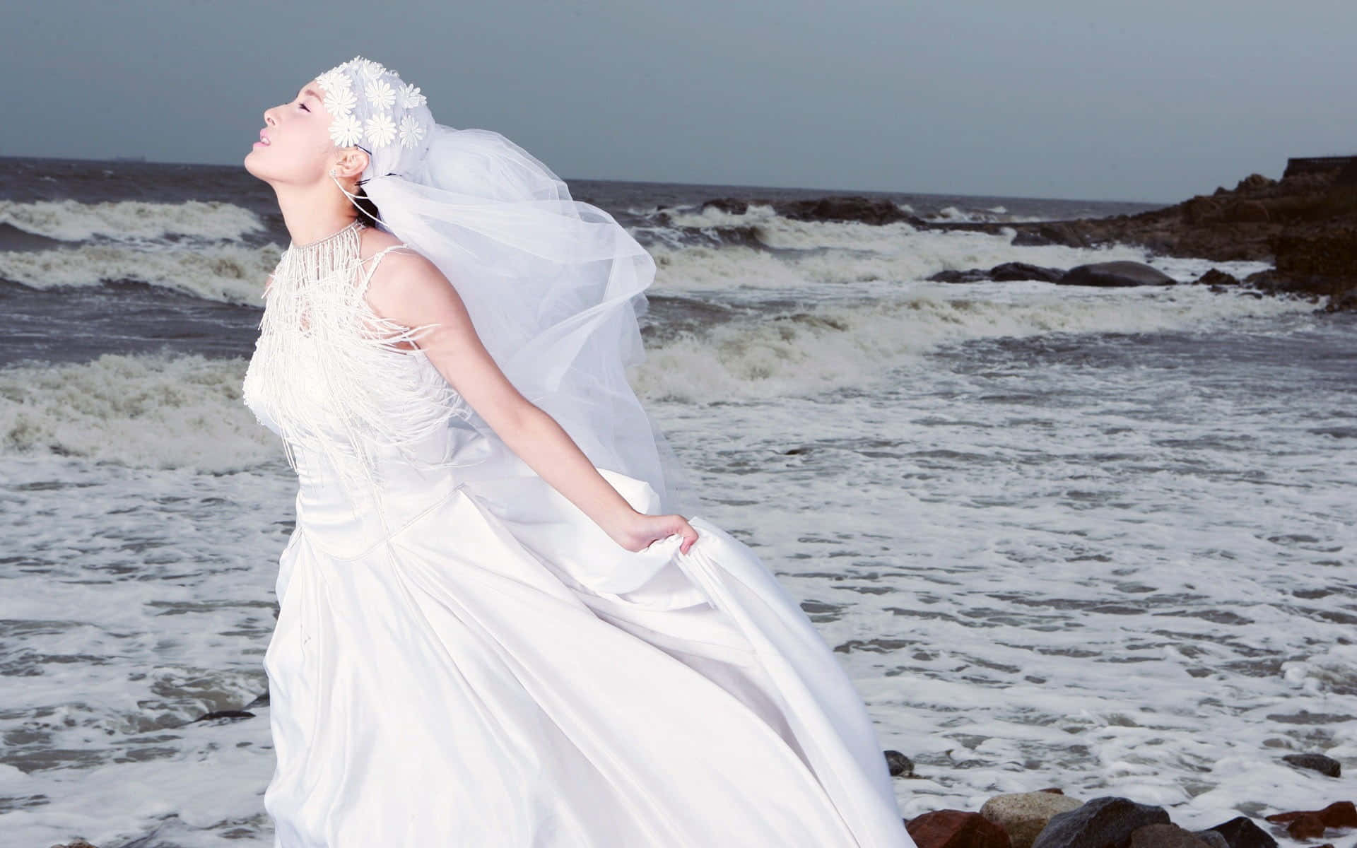 Wedding 桌布 壁紙 — 一位身穿飄逸白色禮服的新娘站在岩石海灘上，海浪拍打著她周圍的水花。