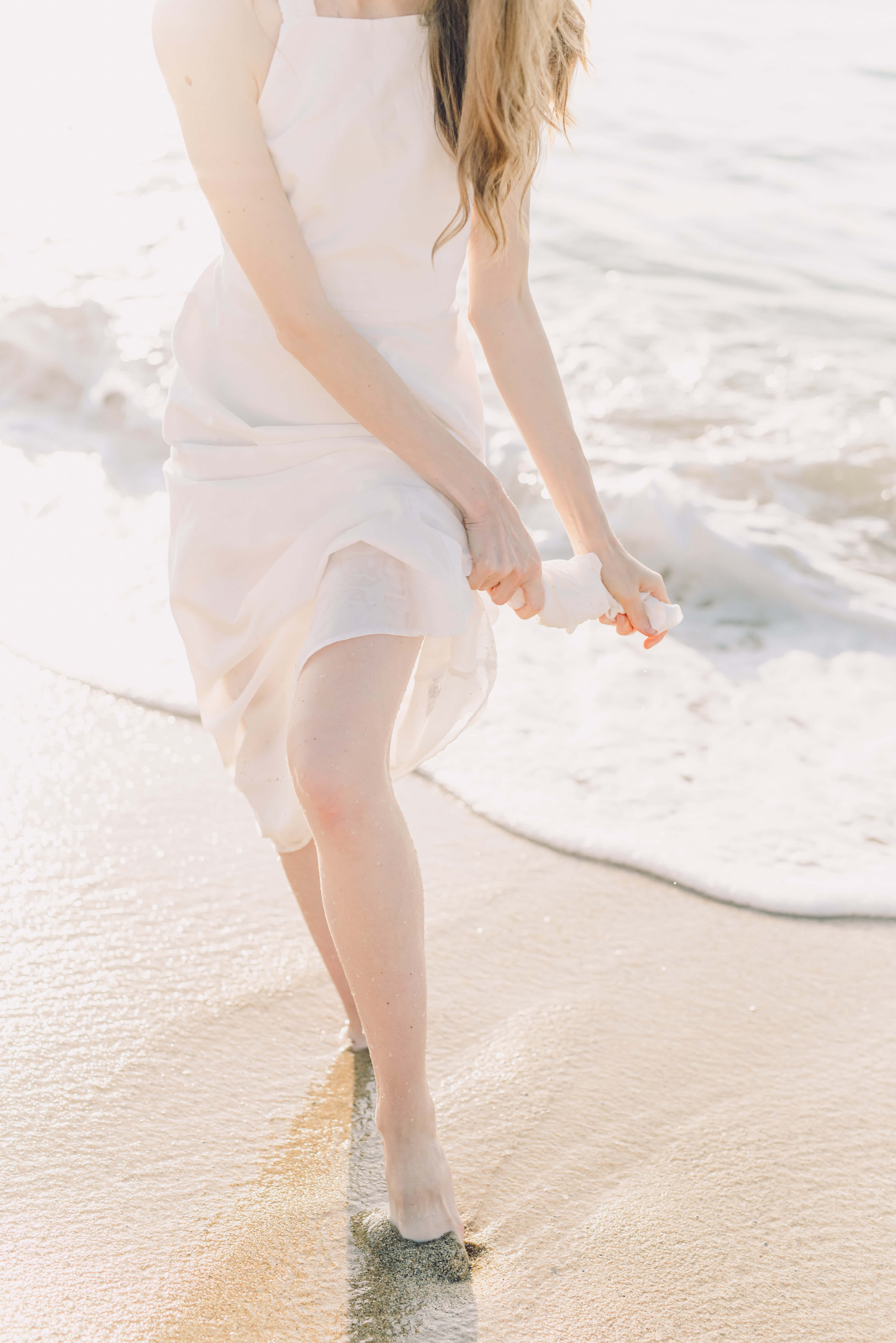 Aesthetic Beachy 桌布 壁紙 — 一位身穿飄逸白色連衣裙的女子赤腳走在陽光照耀的沙灘上，溫柔的海浪輕拍她的雙腳。