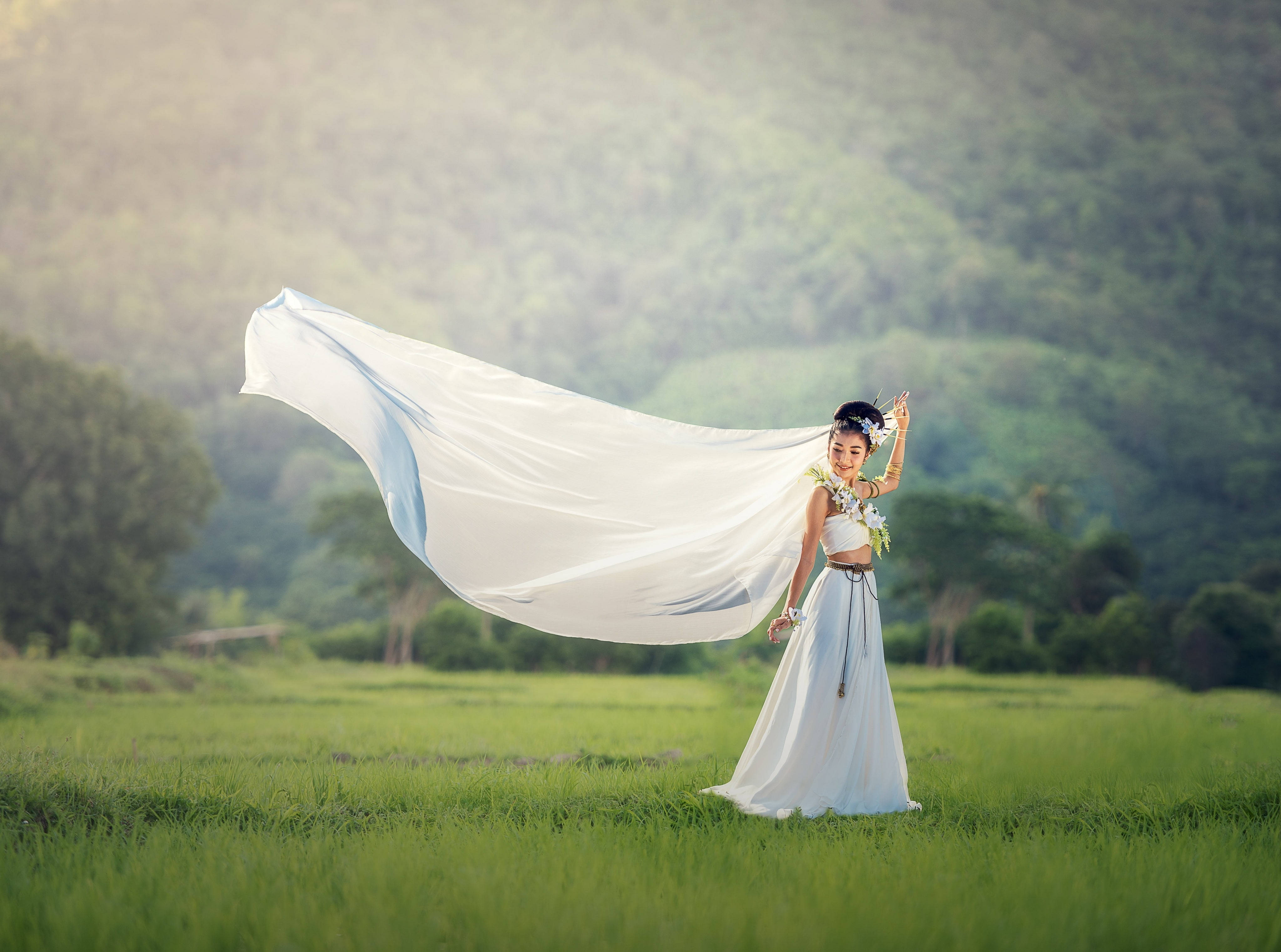 Wedding Dress 桌布 壁紙 — 一位身穿飄逸白色連衣裙的女性，站在陽光明媚的綠色田野中，身後是山脈，她輕薄的布料隨風飄揚。