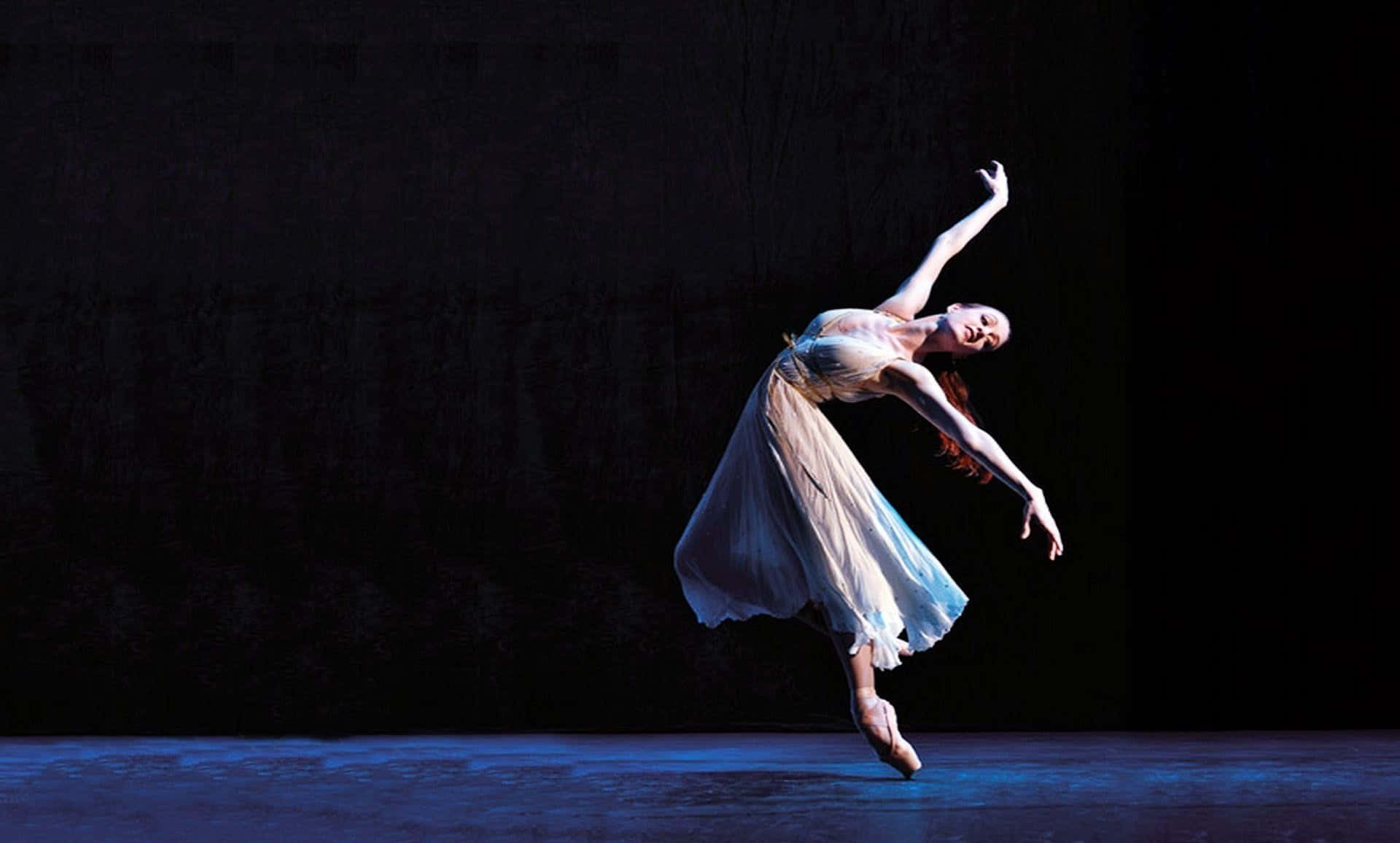 Ballet 桌布 壁紙 — 一位身穿飄逸白色連衣裙的芭蕾舞者在深色背景下，於戲劇性的藍色舞台燈光下優雅地踮腳跳躍。