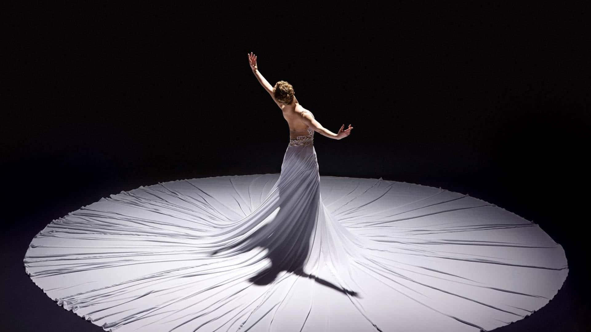 Ballet 桌布 壁紙 — 一位身穿飄逸白色長裙的舞者張開雙臂，布料在黑色背景下的戲劇性聚光燈下向外輻射。