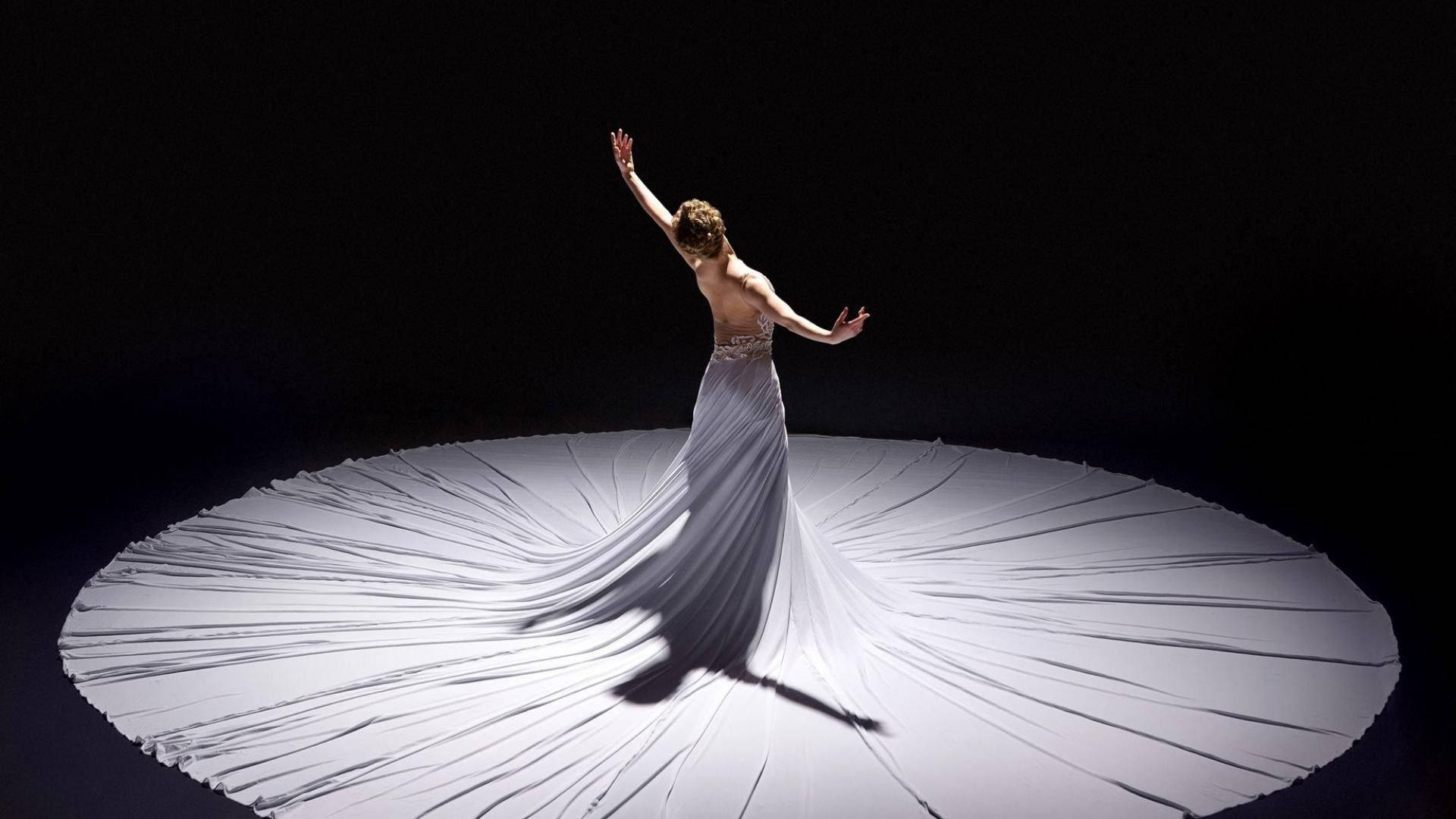 Ballet 桌布 壁紙 — 一位身穿飄逸白色長裙的舞者張開雙臂，布料在戲劇性的聚光燈下向外輻射，映襯著黑色虛空。