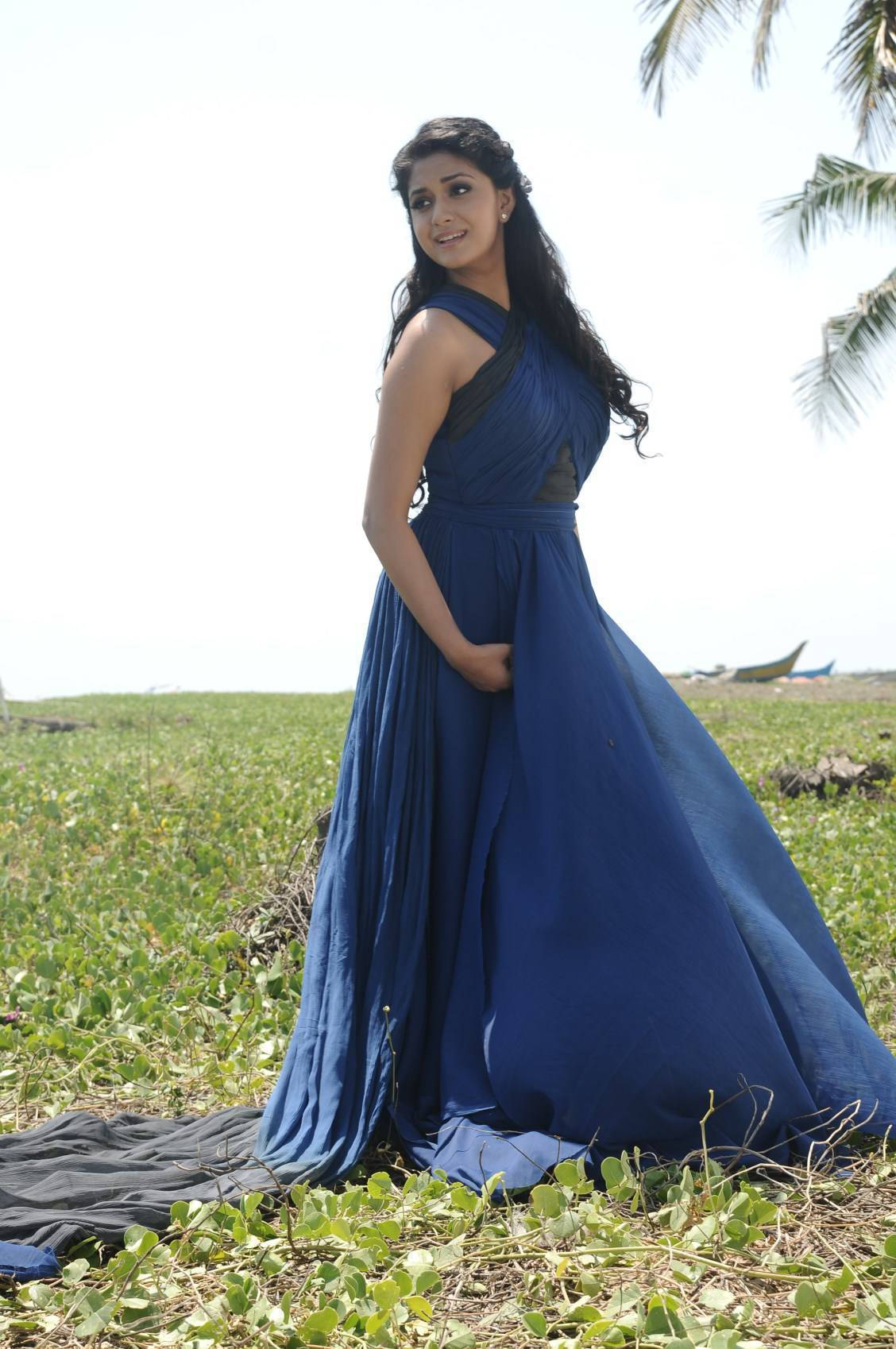 Keerthi Suresh HD 桌布 壁紙 — 一位身穿飄逸皇家藍長袍的女性，優雅地站在陽光明媚的草地上，頭頂棕櫚葉。