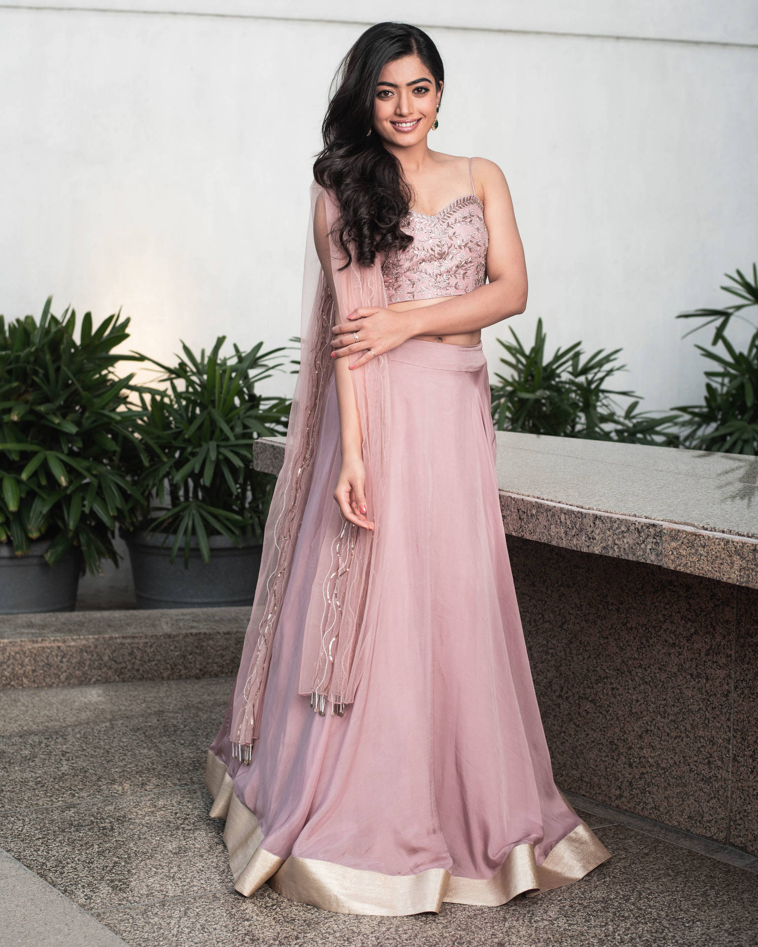 身穿粉紅色 lehenga 的女性在戶外石台和盆栽旁微笑。 — 拉什米卡·曼丹娜 壁紙
