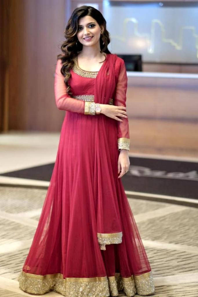 Indian 桌布 壁紙 — 一位身穿飄逸紅色Anarkali套裝、飾有金色刺繡的女性，在現代室內環境中優雅微笑。