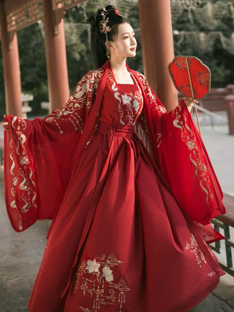 紅色美學動圖 桌布 壁紙 — 一位身穿飄逸紅色傳統長袍、飾有金色刺繡的女子，優雅地站在木亭下，手持相配的扇子。
