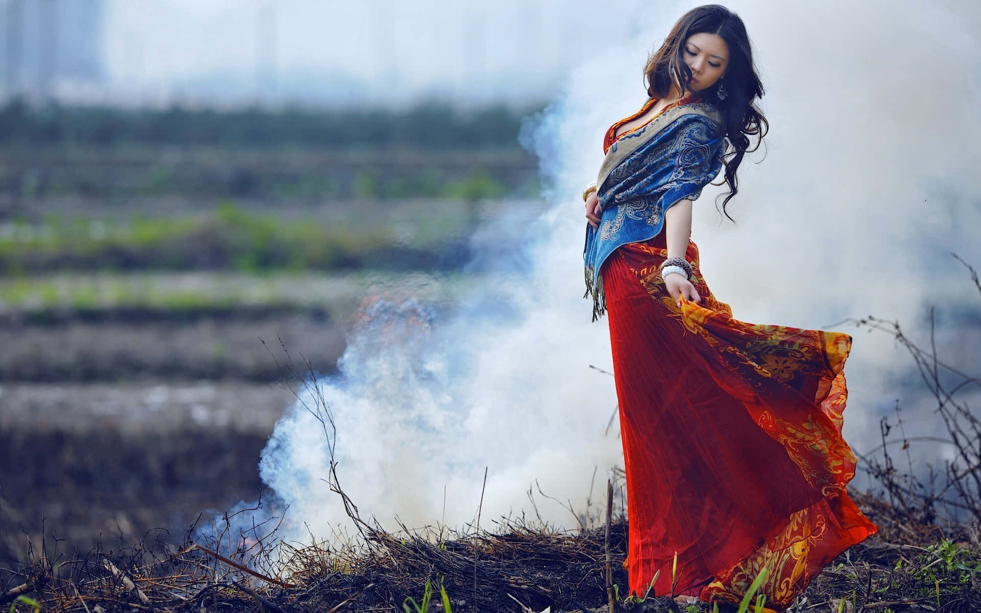 Beautiful Fashion 桌布 壁紙 — 一位身穿飄逸紅藍圖案連衣裙的女子，站在荒蕪田野中翻滾的白煙之中。