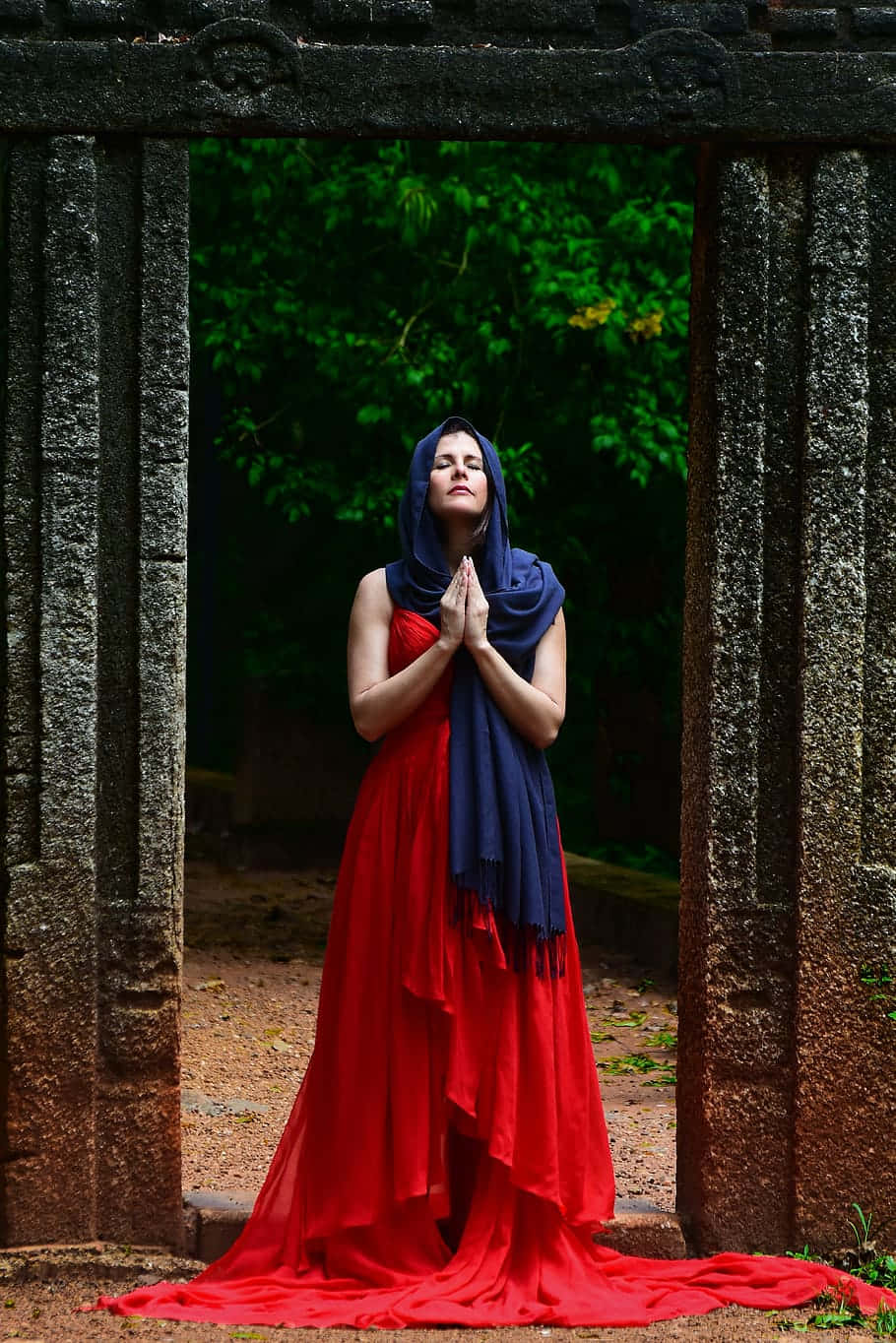 Praying 桌布 壁紙 — 一位身穿飄逸紅裙和藍色頭巾的女性，在一個風化的石拱門內祈禱，周圍環繞著茂密的綠植。